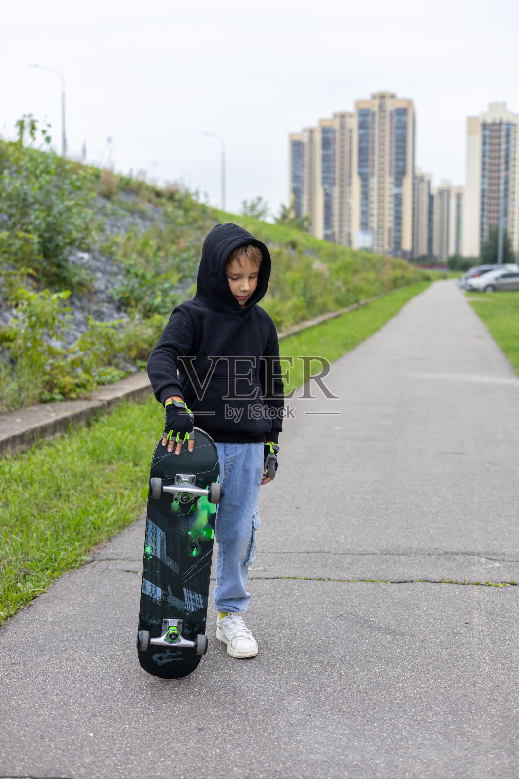 一个时髦的男孩在城市街道上用脚拿着滑板。垂直的。一个孩子在城市里玩滑板的漂亮照片。一个孩子骑滑板的概念。照片摄影图片