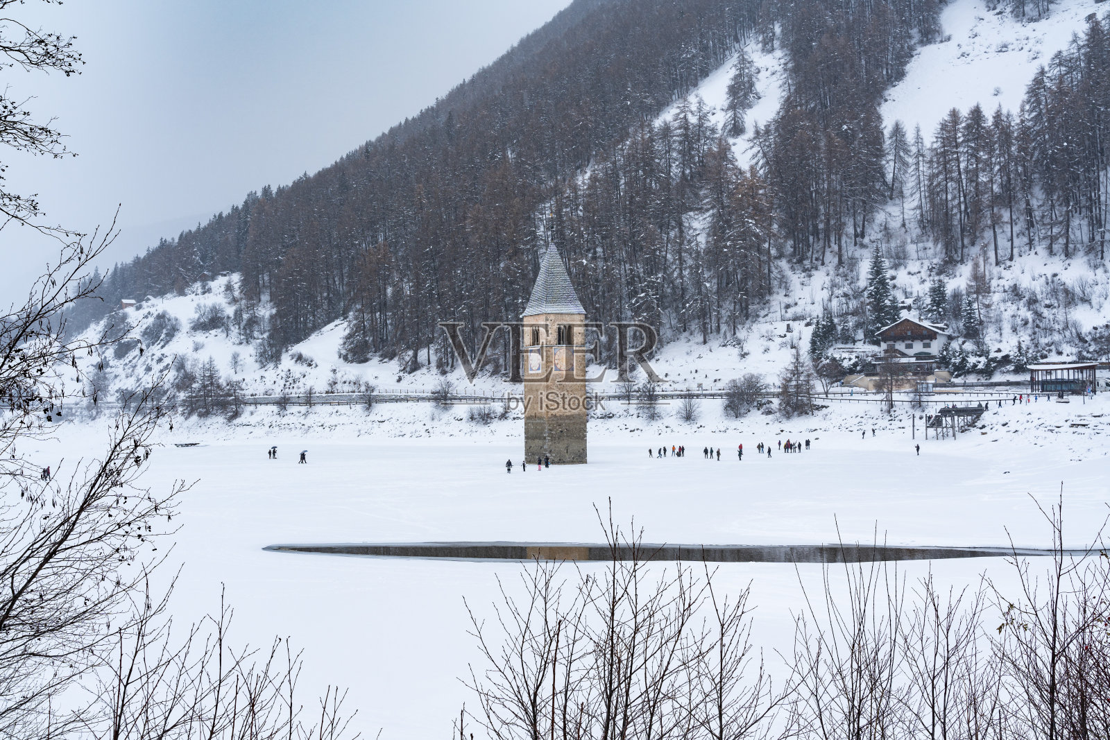 冬天的时候,钟楼矗立在白雪覆盖的雷西亚湖上,背景是树木覆盖的山脉照片摄影图片