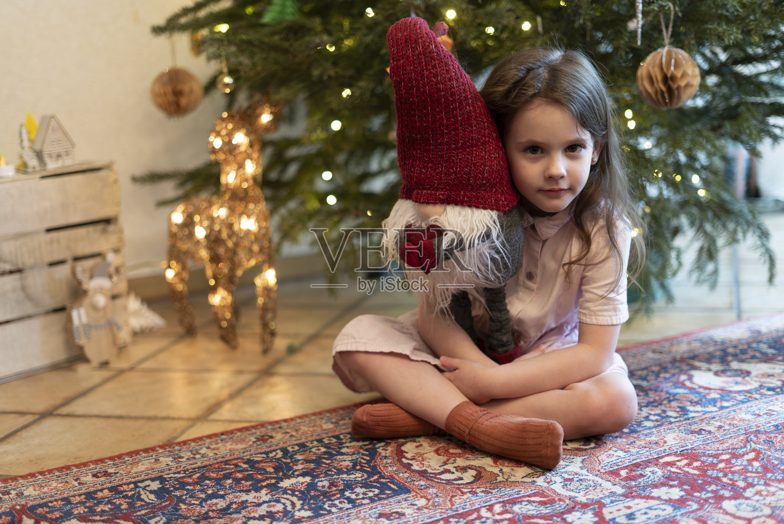 一个悲伤的美丽小女孩穿着盛装坐在圣诞树下,抱着一个圣诞小精灵。一只戴着花环的鹿站在附近照片摄影图片