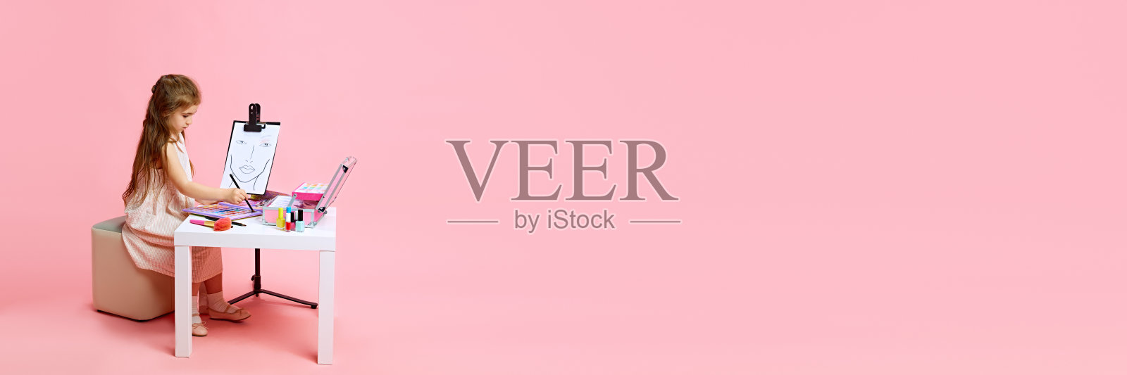 横幅。女孩梦想成为化妆师,用各种化妆品在淡粉色的背景下练习脸部素描。照片摄影图片