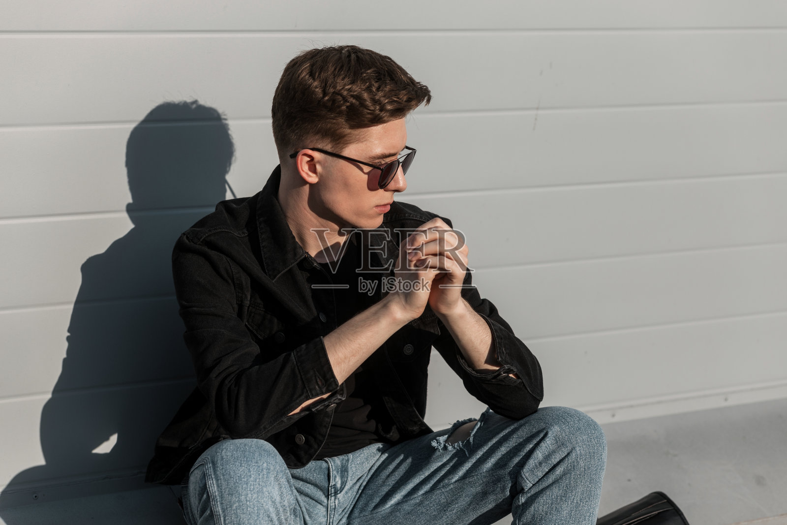 酷酷的美国年轻潮人戴着时髦的太阳镜,穿着时髦的休闲牛仔衣服,发型,在阳光明媚的日子里在街上的建筑物附近休息。现代男士时尚模特在城市。青春的风格。照片摄影图片