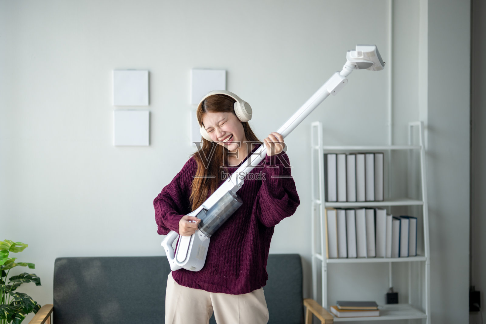 一个女人在像弹吉他一样弹吸尘器。她戴着耳机,面带微笑。房间里有一张沙发和一个书架照片摄影图片