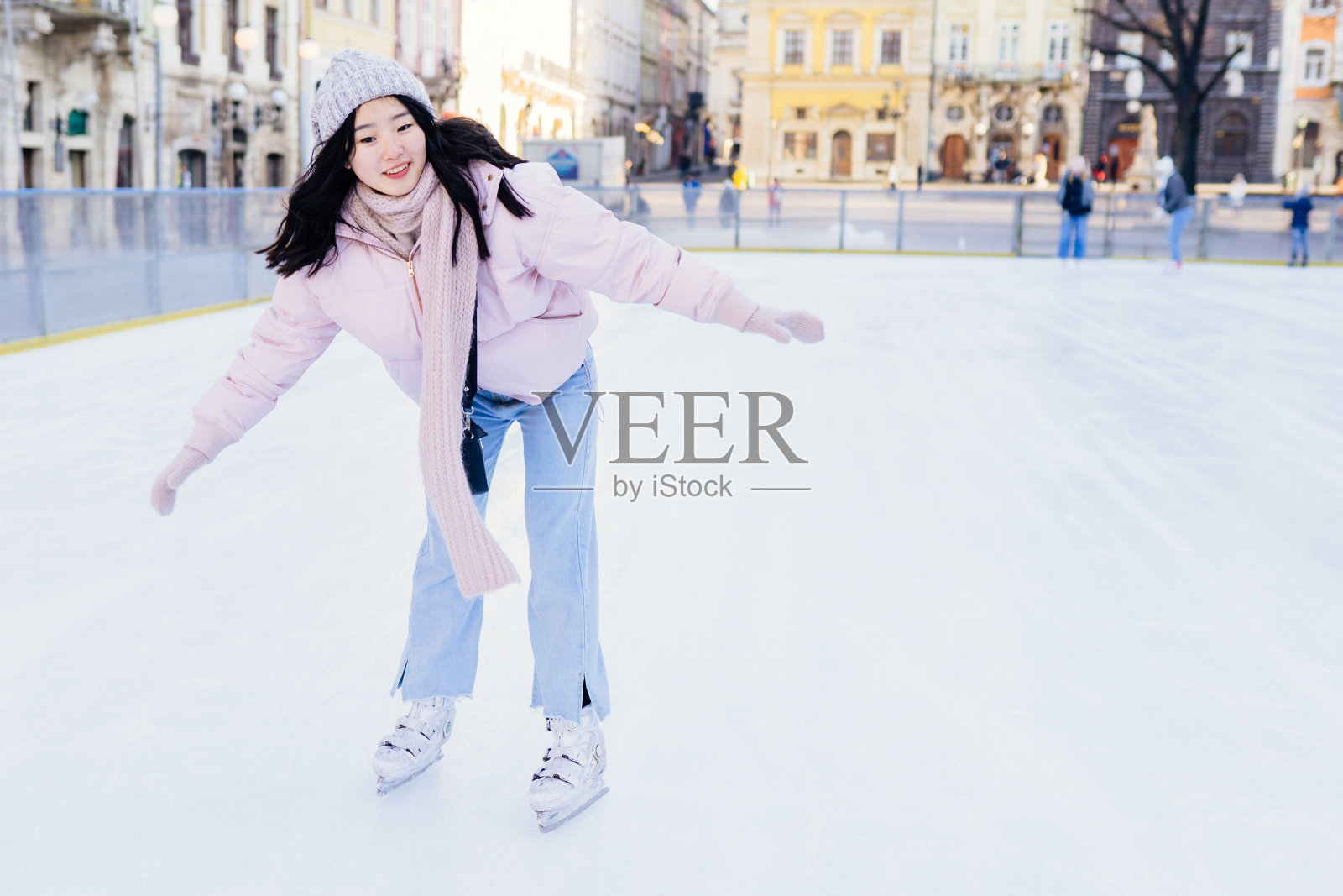 亚洲女子在户外滑冰。韩国女性穿着粉粉色的夹克,戴着暖和的针织帽、围巾和手套。健康的生活方式,城市广场的运动理念。冬天的天气晴朗。全身照片摄影图片