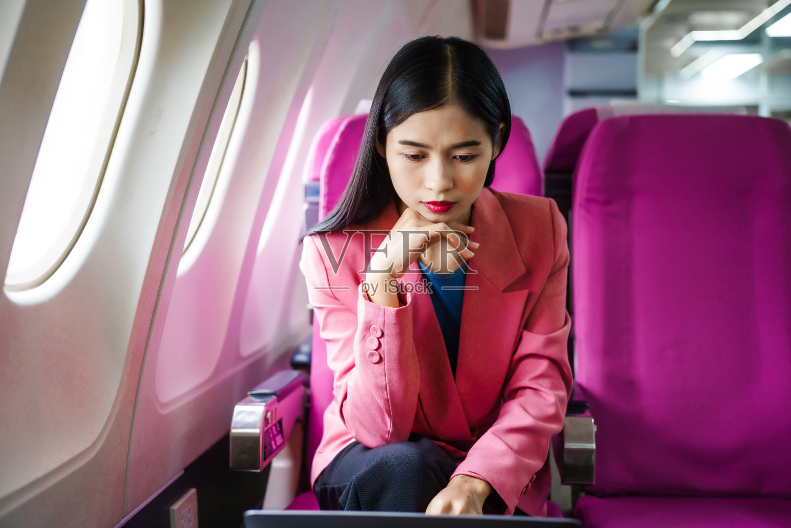 一位年轻的亚洲女乘客在飞行过程中坐在窗口,拿着登机牌,头顶上放着随身行李,系着安全带,享受着旅程。照片摄影图片