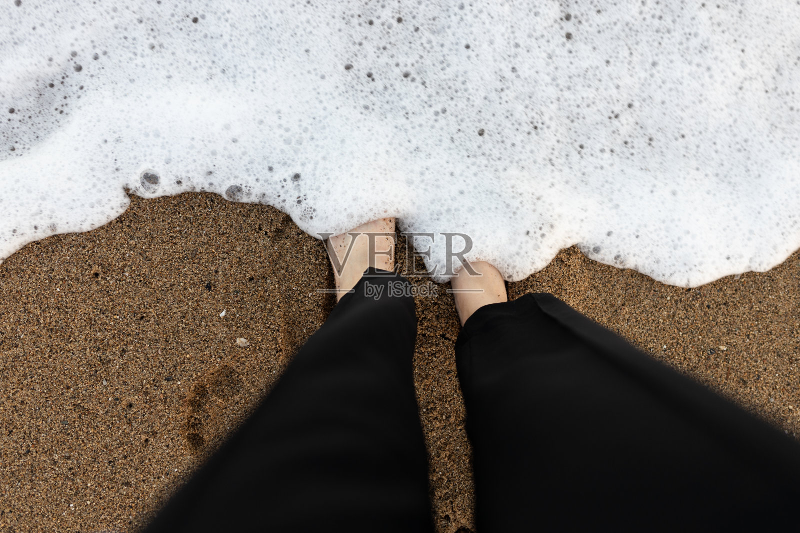 第一人称视角。脚踩在沙滩背景上。俯视图在沙滩上的腿与波浪运动来的脚,发泡的海洋纹理。暑假概念。赤脚站在海边自拍的女人照片摄影图片