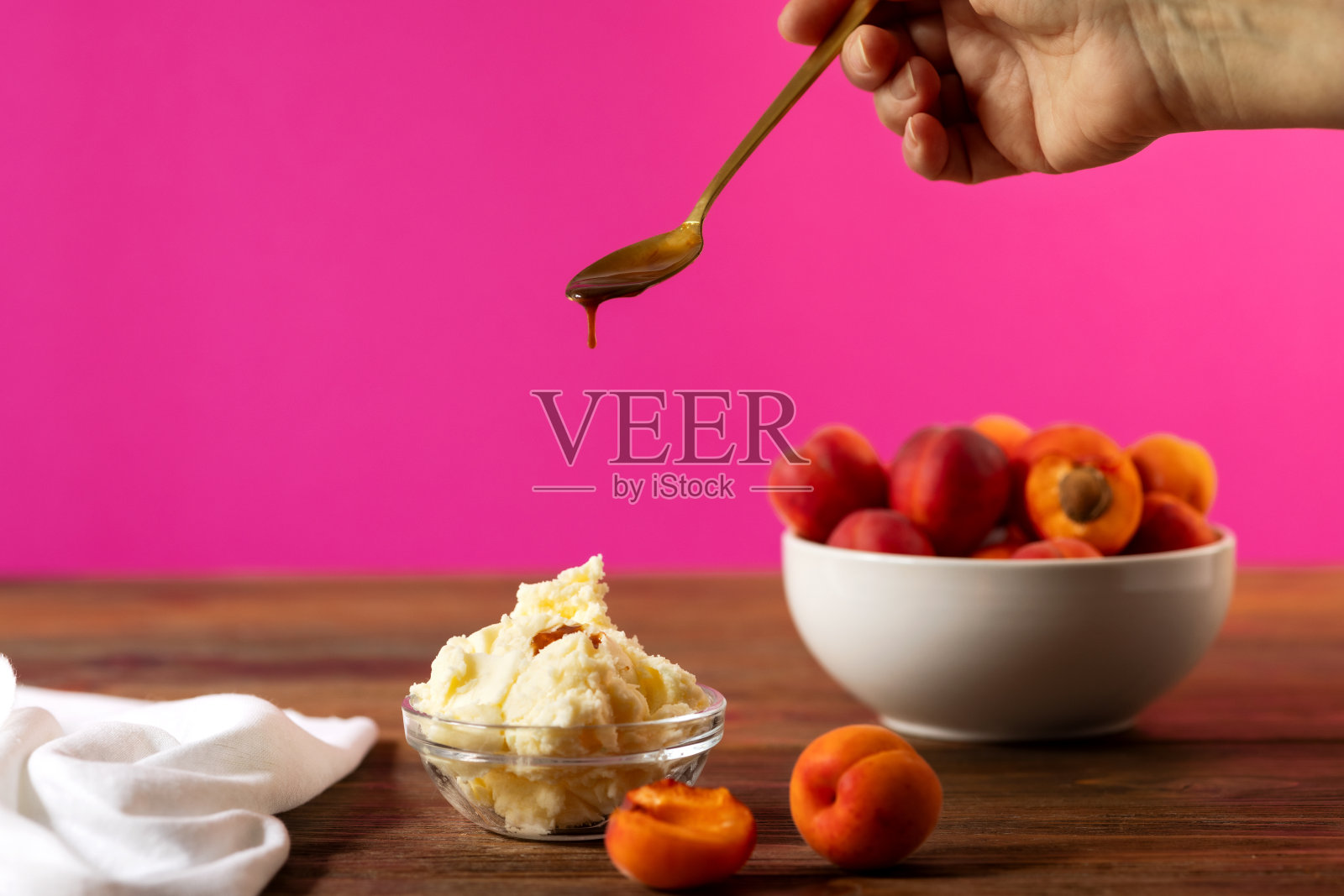 女人的手用勺子将焦糖冰淇淋倒入玻璃碗中。木桌上的白碗和杏子。照片摄影图片