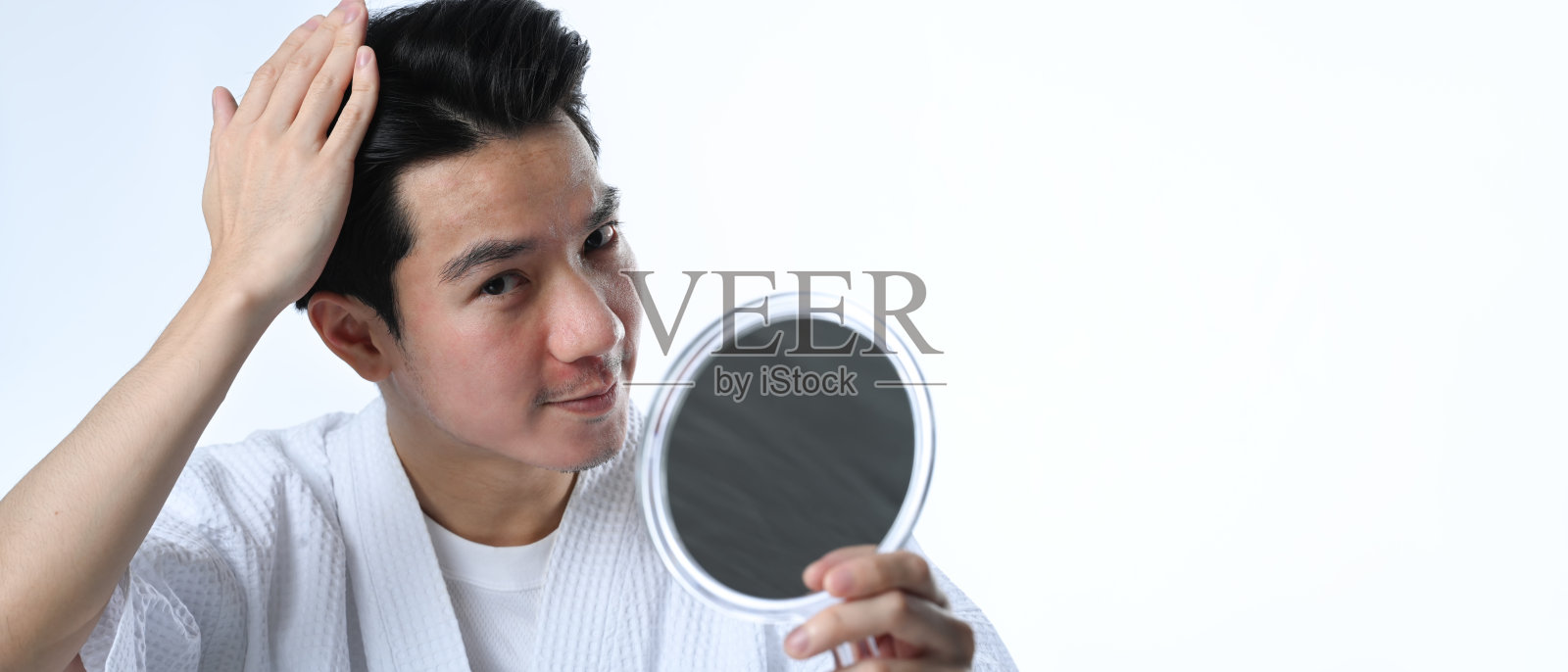 年轻的亚洲男子一边审视自己的头发,一边看着白色背景的镜子照片摄影图片