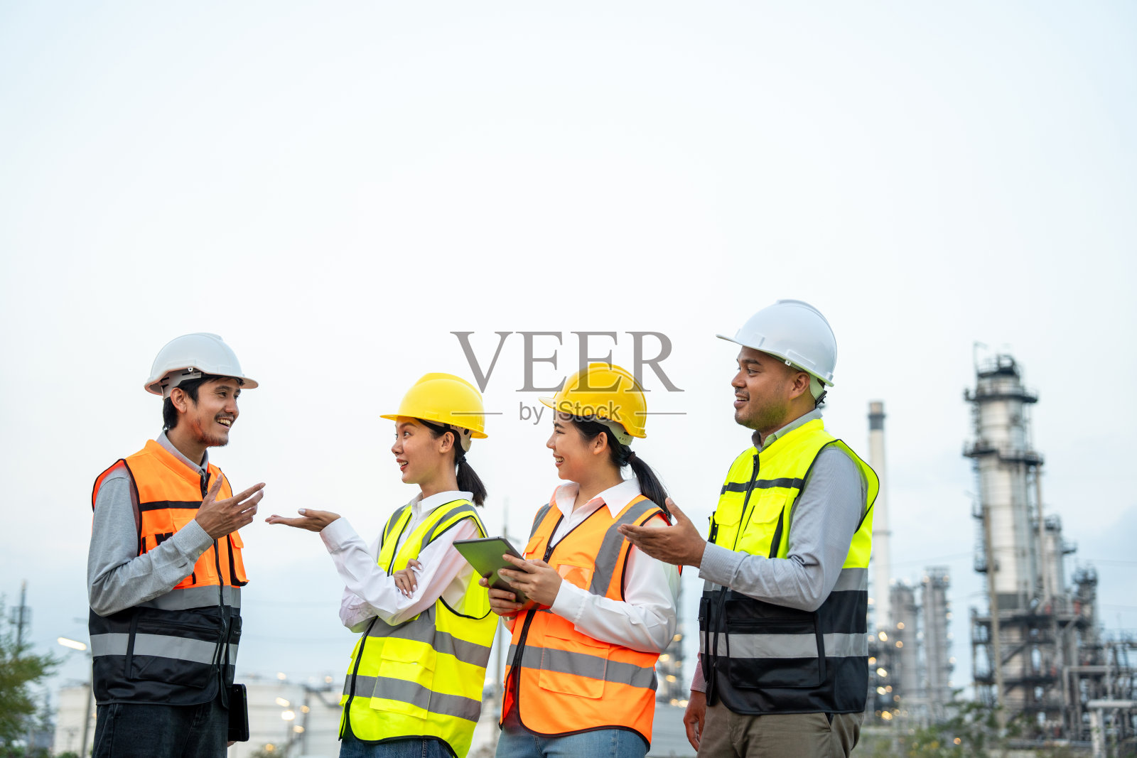 一群戴着安全帽的亚洲工程师站在炼油厂前。工业园区天然气石化。工厂储油罐和管道。工人们在炼油厂建设中进行团队合作照片摄影图片