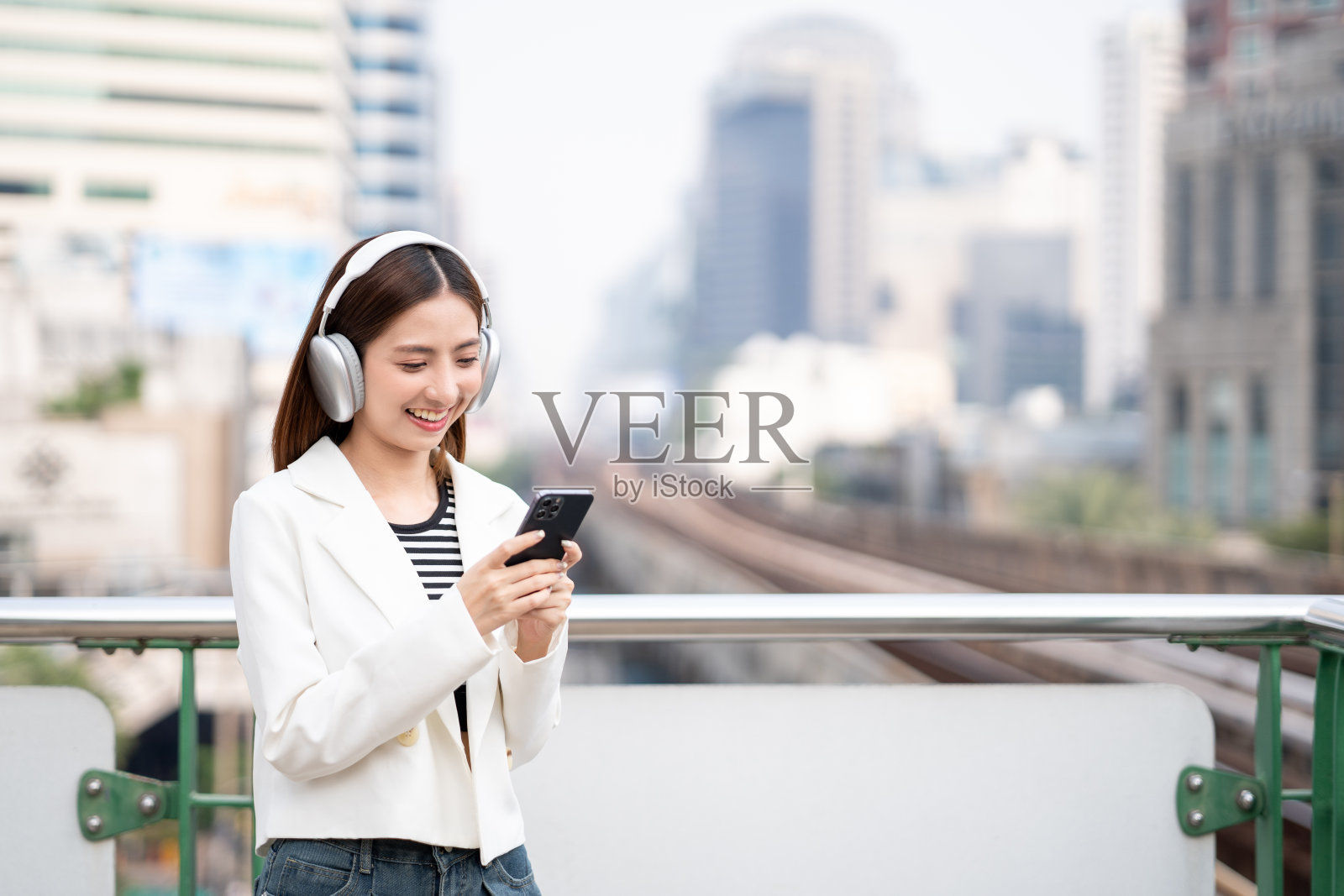 年轻的亚洲女性在城市中行走时使用智能手机。快乐美丽的亚洲女性,戴着耳机,站在城市办公大楼用手机听音乐。生活方式的人照片摄影图片