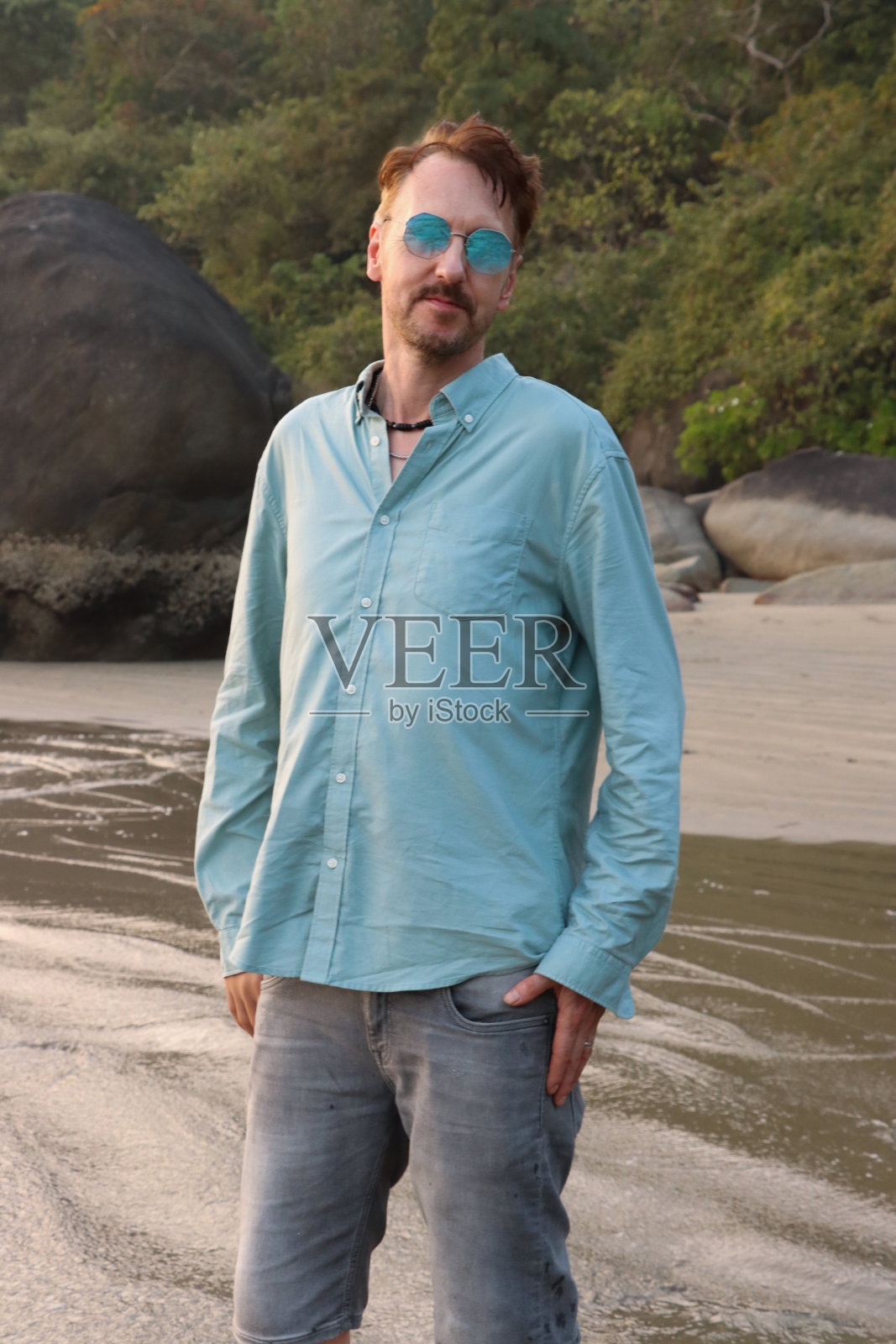 广角图像的人站在热带海滩,海边,戴着镜面蓝色太阳镜和蓝色衬衫,附近的海岸日落。照片摄影图片