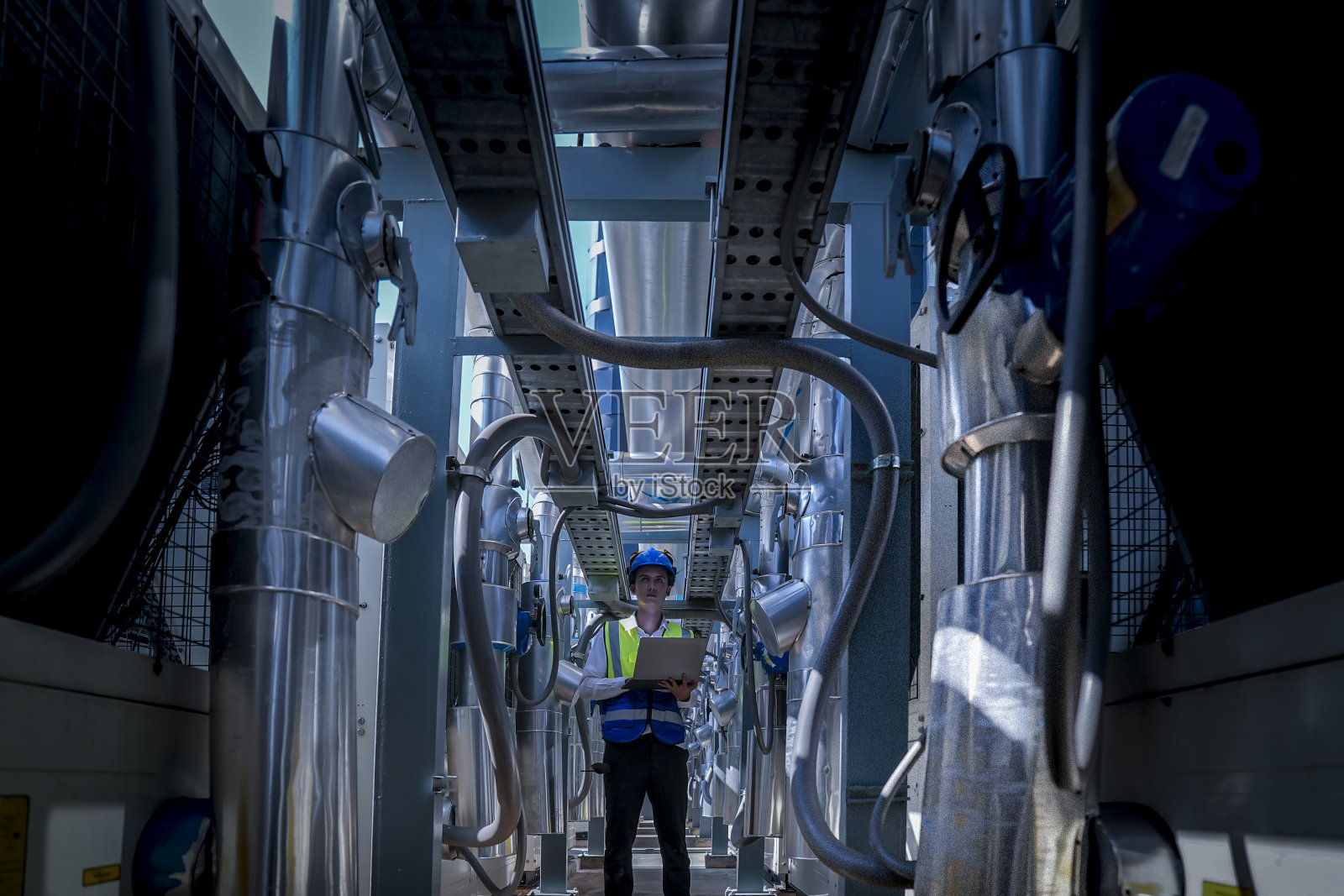 一名技术人员穿着明亮的安全背心和头盔,对复杂的工业设备进行彻底检查。该场景突出了工程任务中精度和安全性的重要性。照片摄影图片