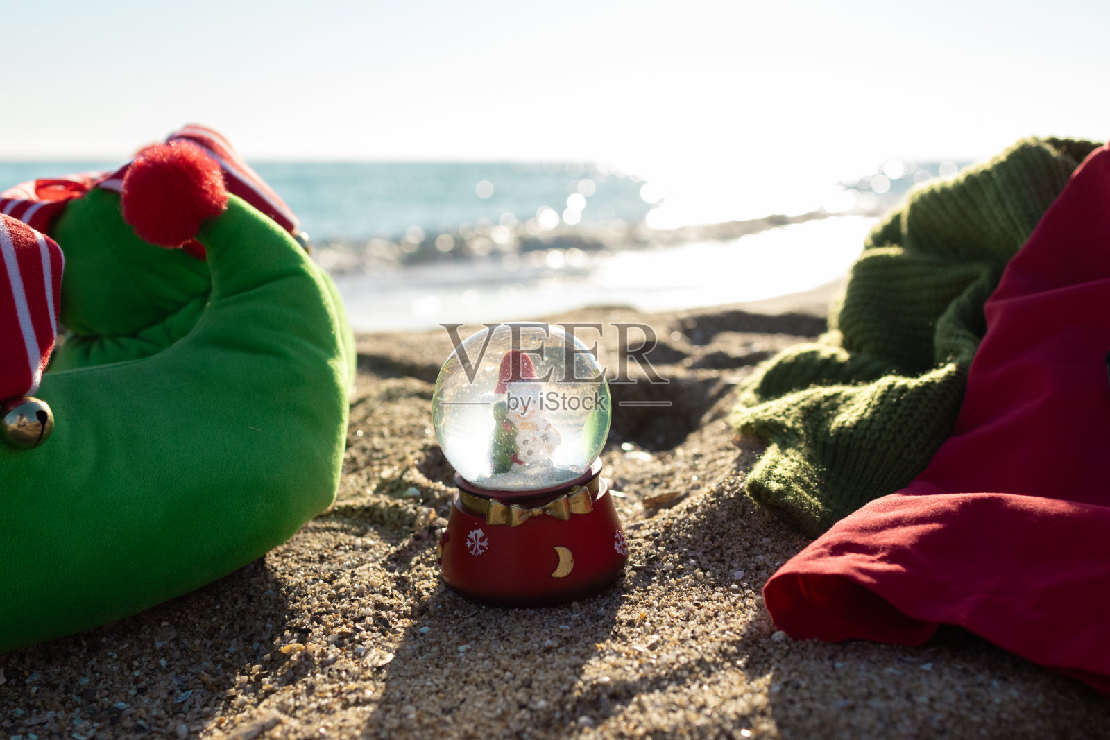 圣诞装饰水晶球里面有雪花和水在沙滩前的沙滩上。圣诞精灵服装和拖鞋。照片摄影图片