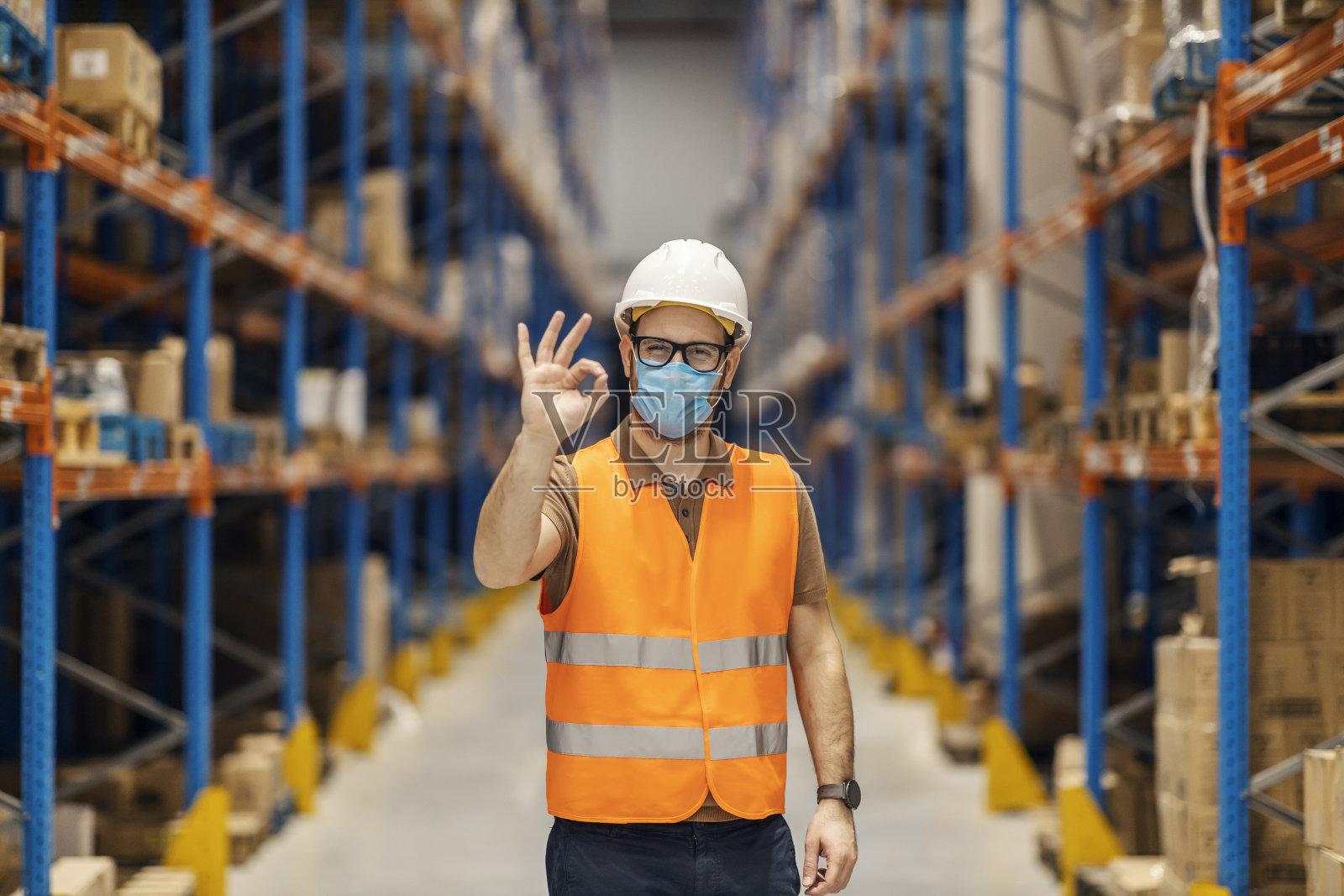 大流行期间,一名仓库工人戴着口罩站在设施里,做着ok的手势。照片摄影图片