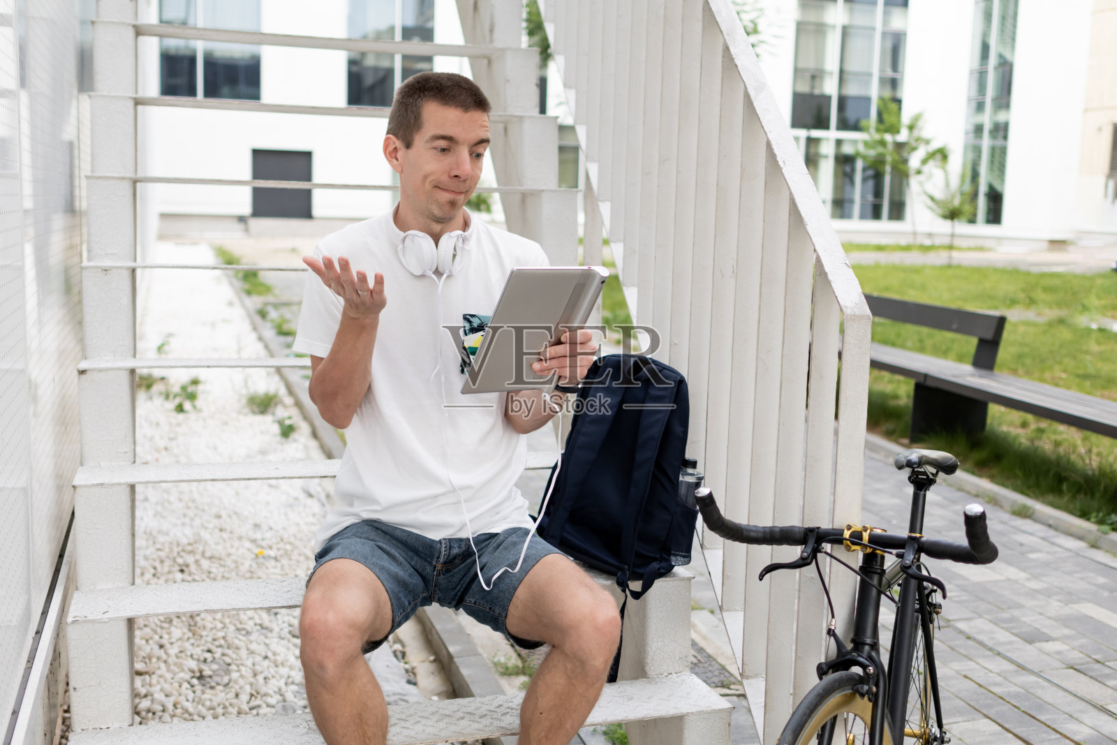 戴着耳机的快乐放松的中年男子正在他的数码平板电脑上进行视频通话。可持续的自行车旅行。在现代住宅区休息一下。照片摄影图片