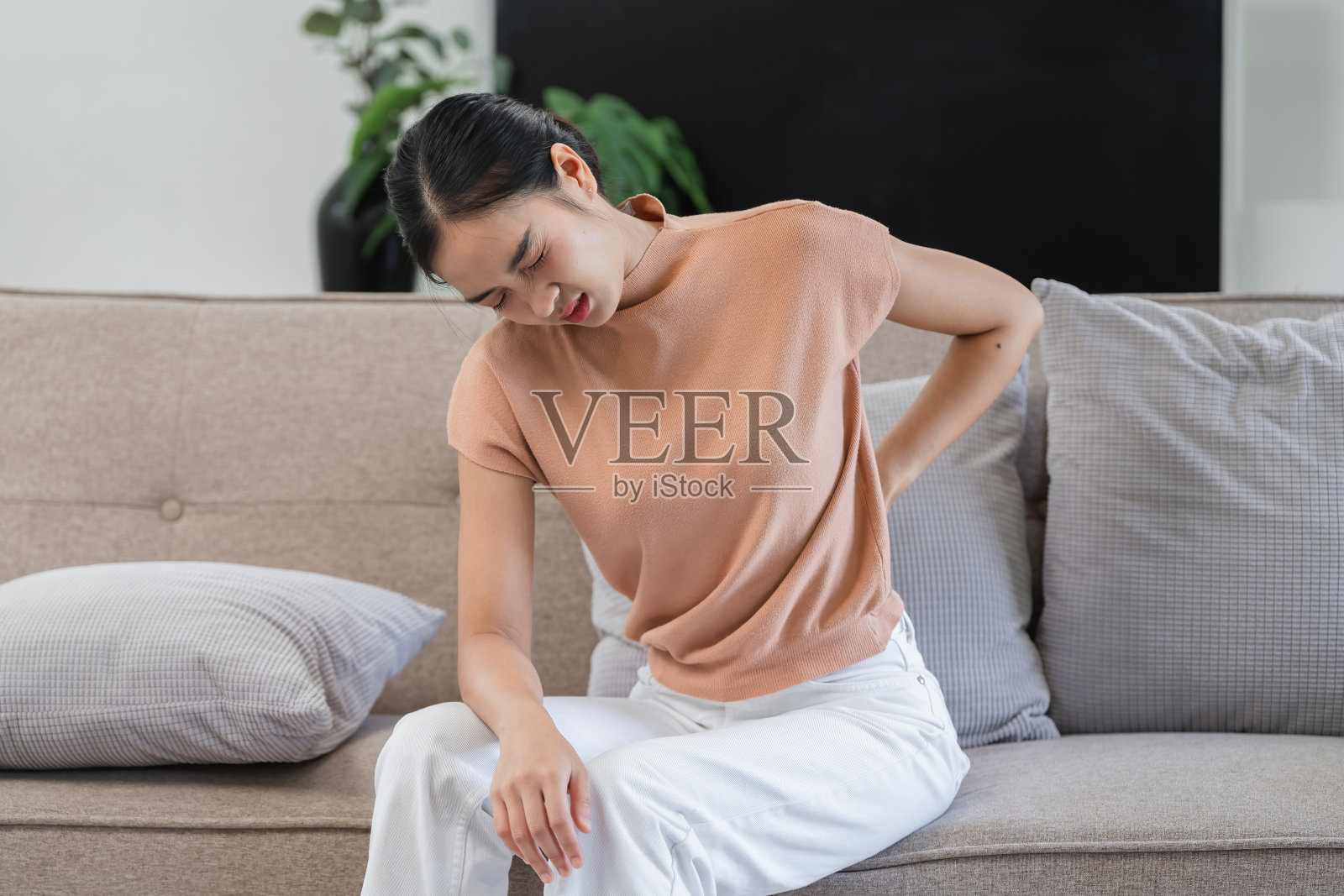 年轻女子坐在明亮的现代客厅的沙发上,有绿色植物和自然光,正在经历背部疼痛照片摄影图片