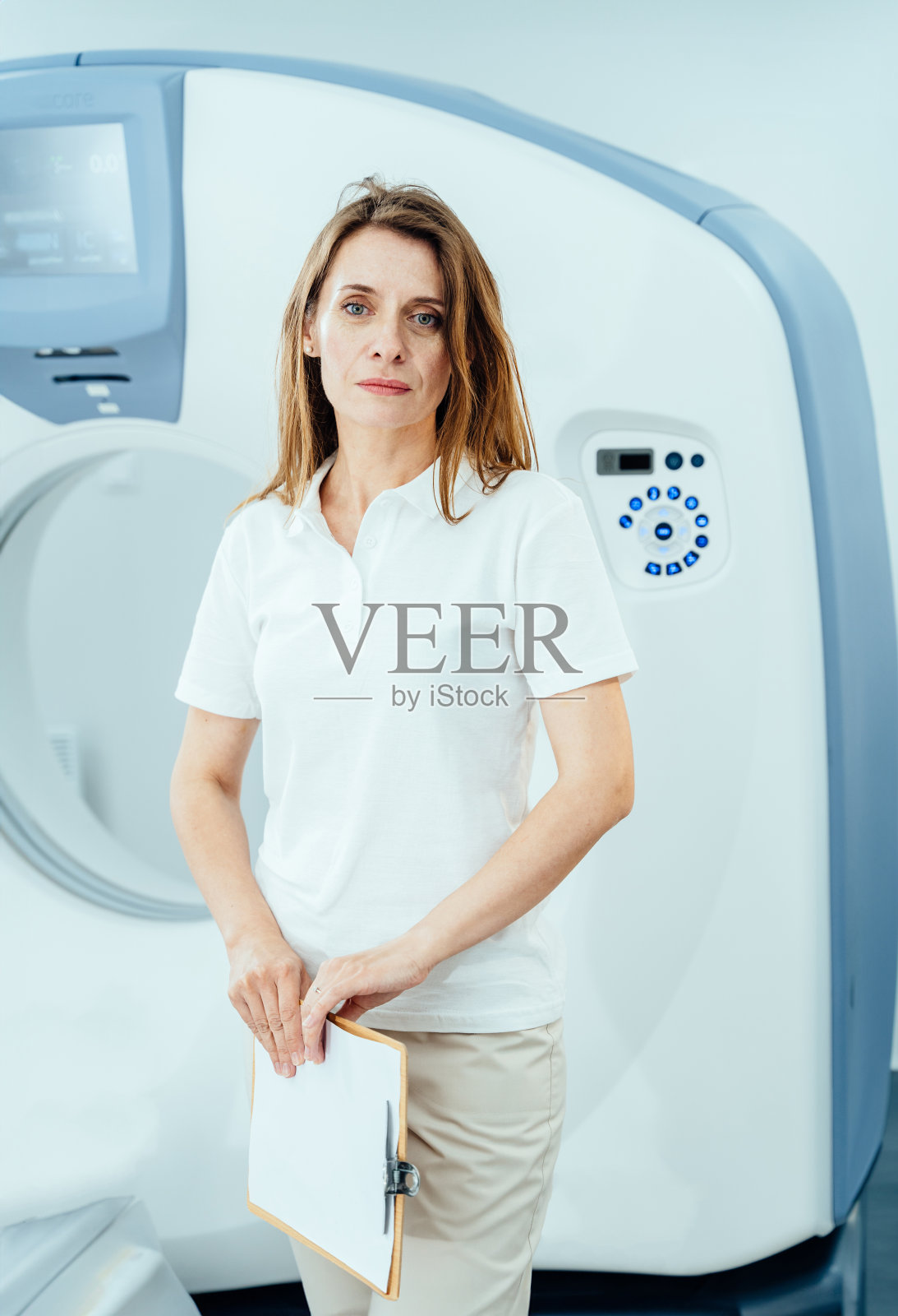 一位女士站在GE核磁共振成像仪前。她穿着一件白衬衫,拿着一个剪贴板照片摄影图片