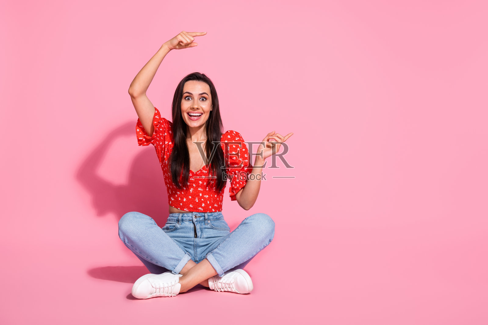 漂亮的年轻女士的全身肖像直接手指空的空间,坐在地板上,穿在粉红色的背景上孤立的顶部照片摄影图片