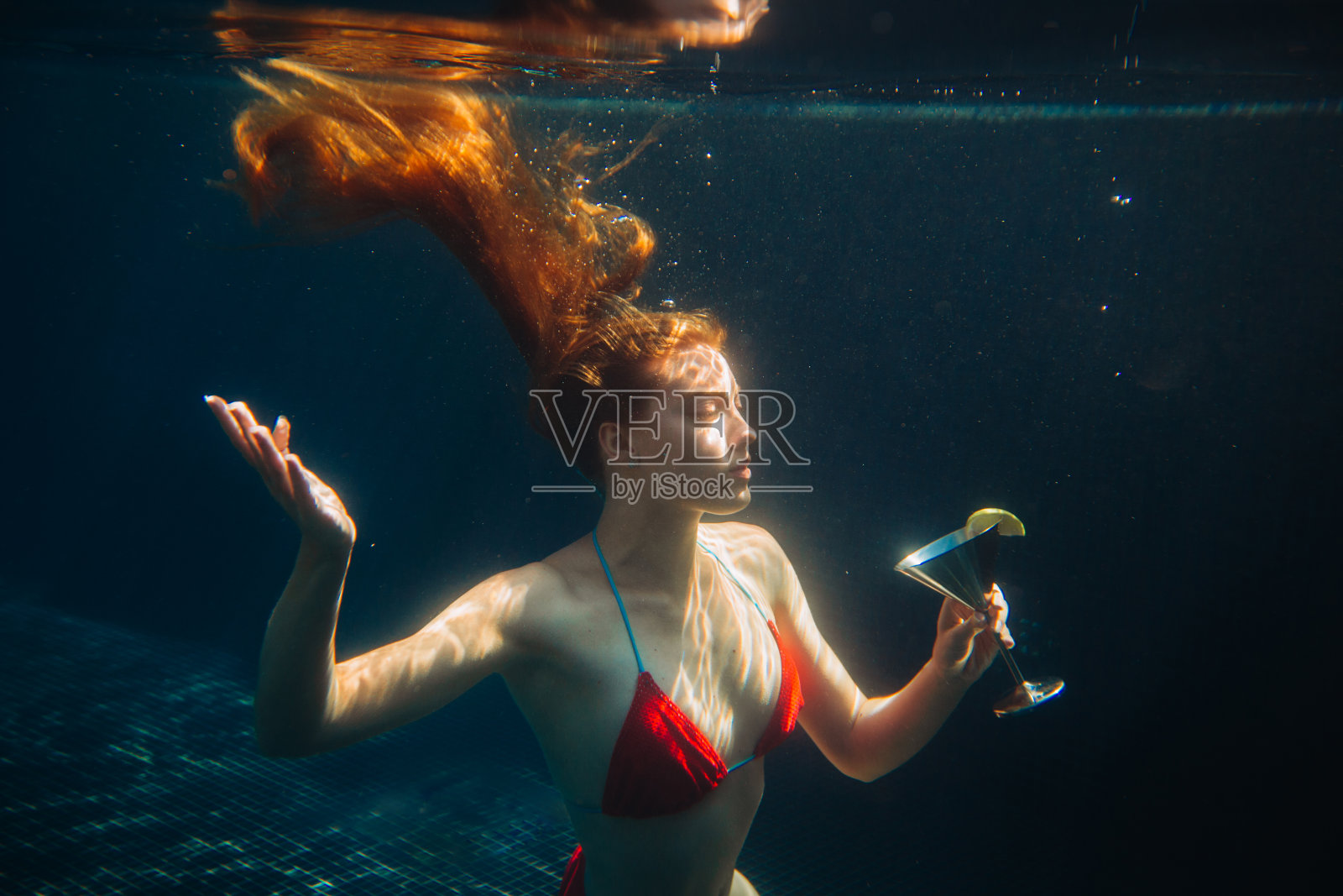 美丽的红发女人在游泳池里拿着鸡尾酒摆姿势。暑假概念。照片摄影图片