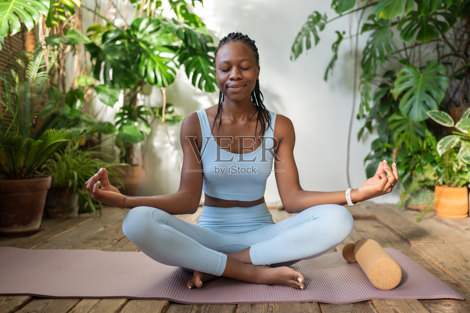 满意的黑人妇女闭着眼睛,坐成莲花式,在热带室内植物周围沉思。照片摄影图片