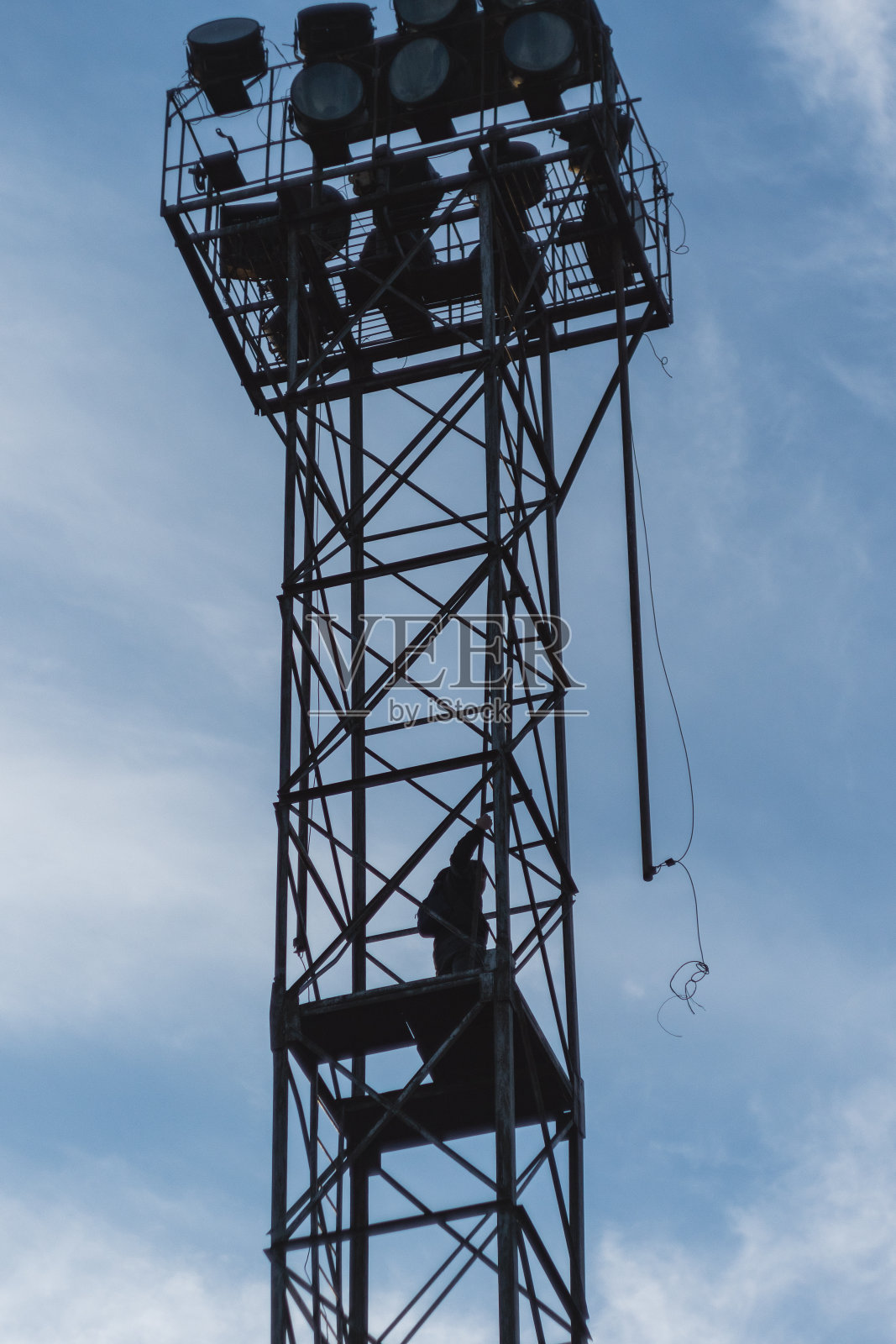 在天空的映衬下,可以看到一个工人的剪影,他正在一座高高的金属塔上工作。在塔的顶部,由金属桁架组成,有几个信号灯。照片摄影图片