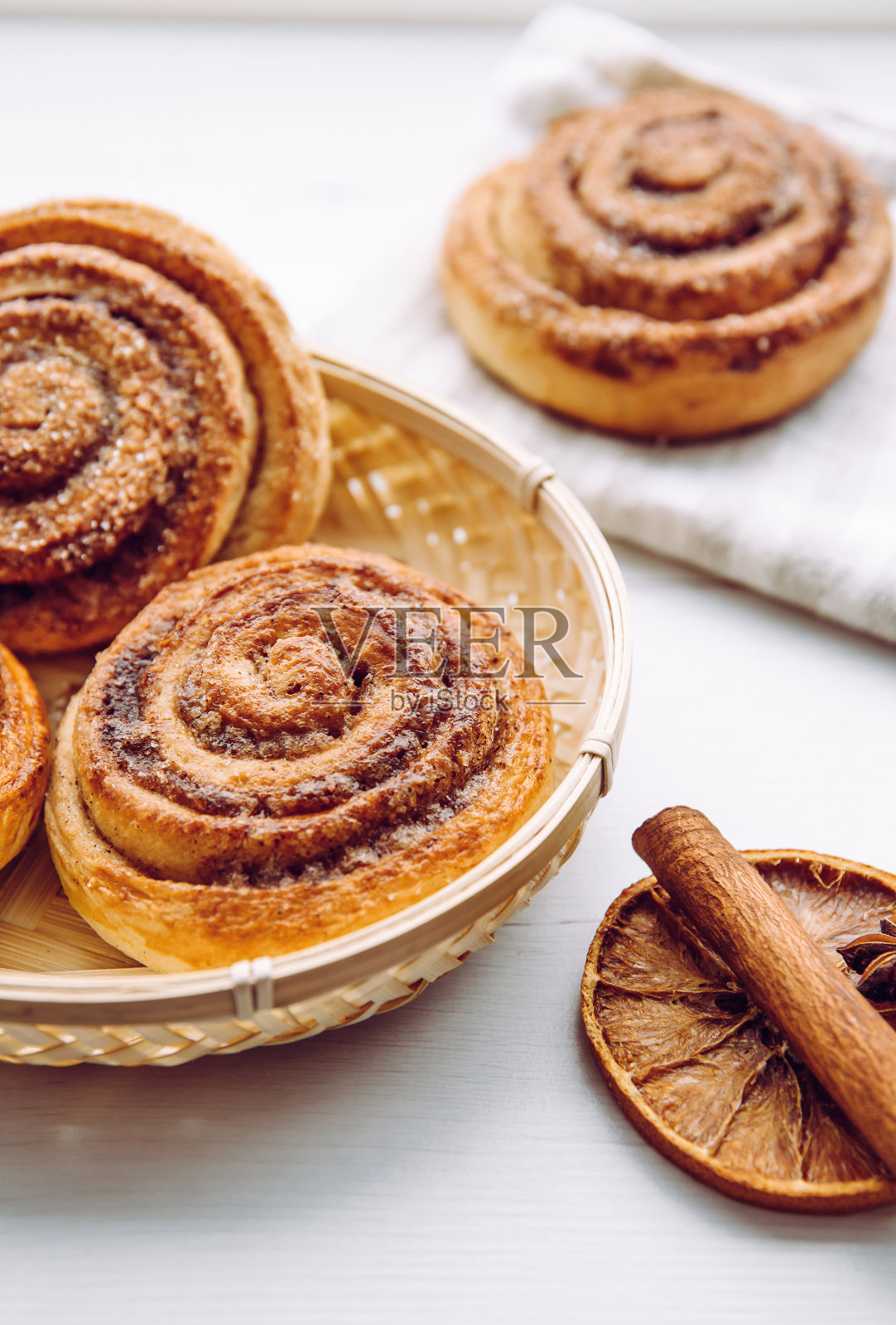 在家庭厨房的白色餐桌上,新鲜出炉的自制肉桂卷放在篮子里,肉桂棒作为装饰。选择性对焦,侧视图,自然背光。照片摄影图片