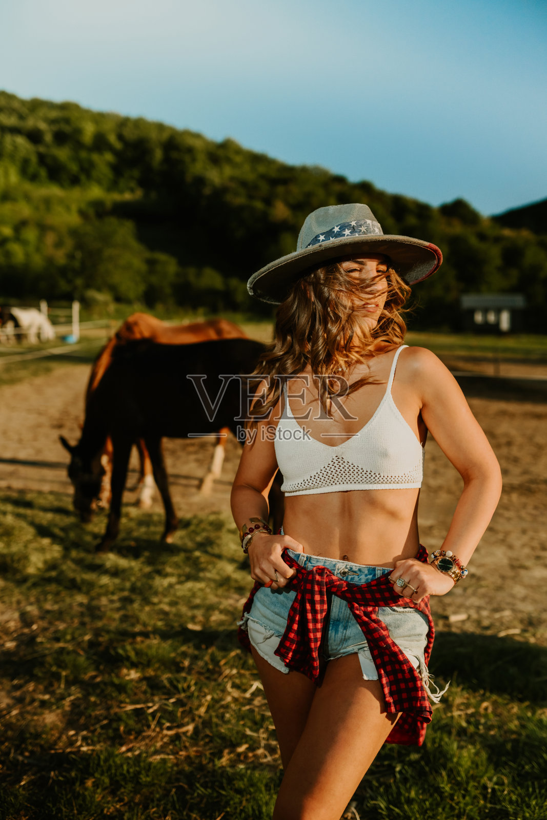 年轻诱人的牧场主女孩在牧场上与吃草的马合影。照片摄影图片