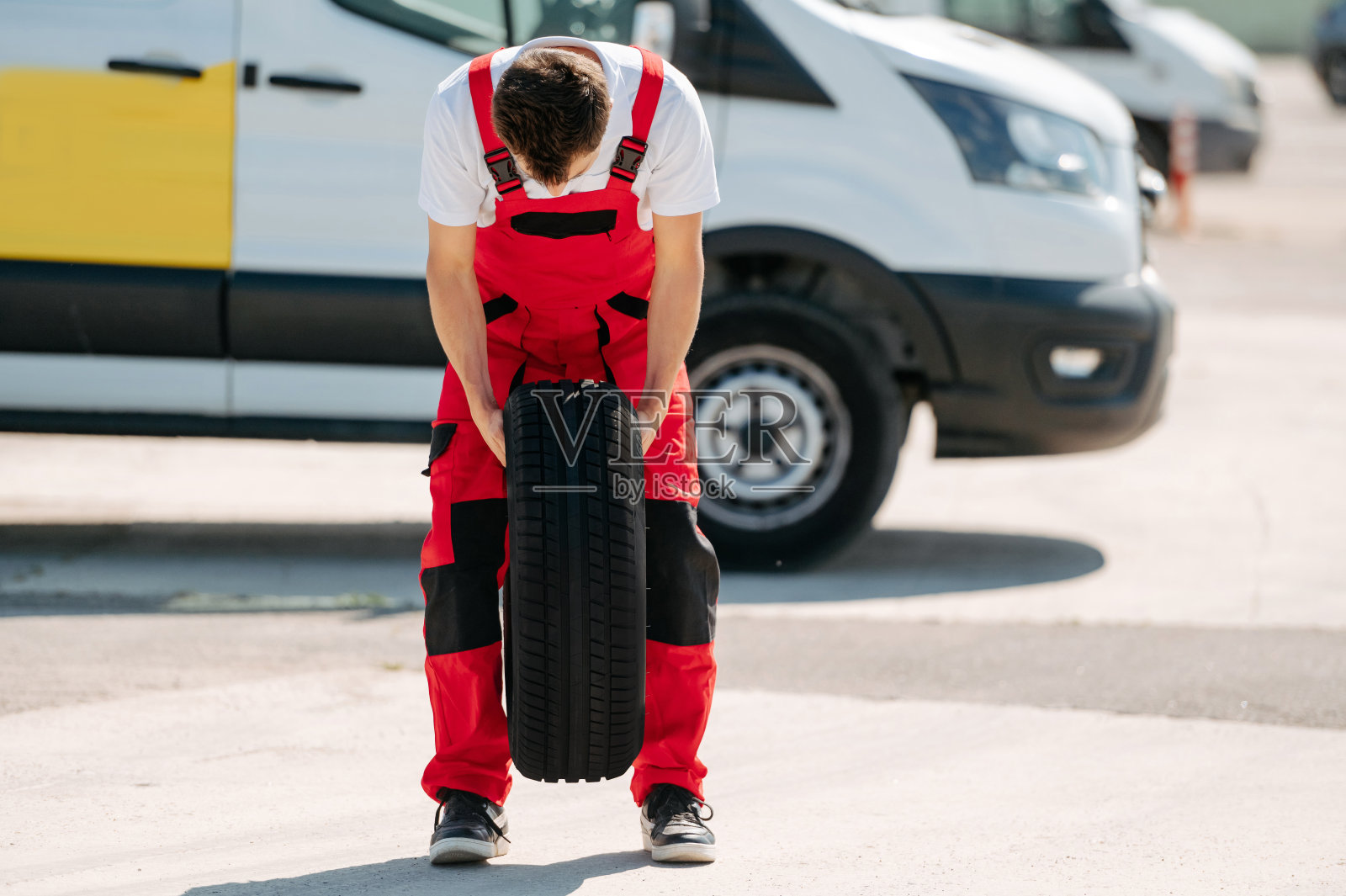 年轻的微笑男子,穿着红色工作服,汽车修理工拿着汽车轮胎照片摄影图片
