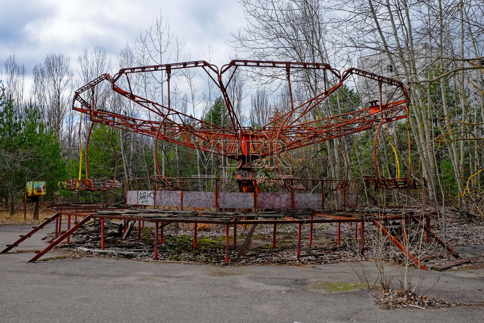 生锈的废弃旋转木马被杂草丛生的树木和破旧的建筑包围。照片摄影图片
