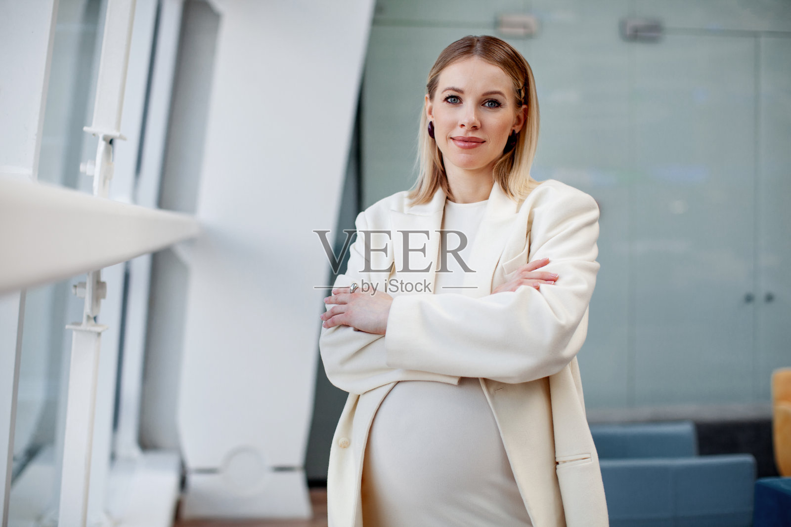 美丽,自信,怀孕的金发女商人,穿着时尚时尚的衣服,白色连衣裙和运动夹克,在蓝色背景的公司办公室里看着镜头照片摄影图片