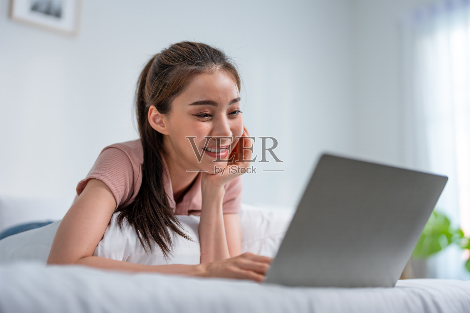 女青年早上在家的床上用笔记本电脑聊天。快乐随意的漂亮女人躺在卧室享受假期周末用笔记本电脑上网与技术在家里交流照片摄影图片