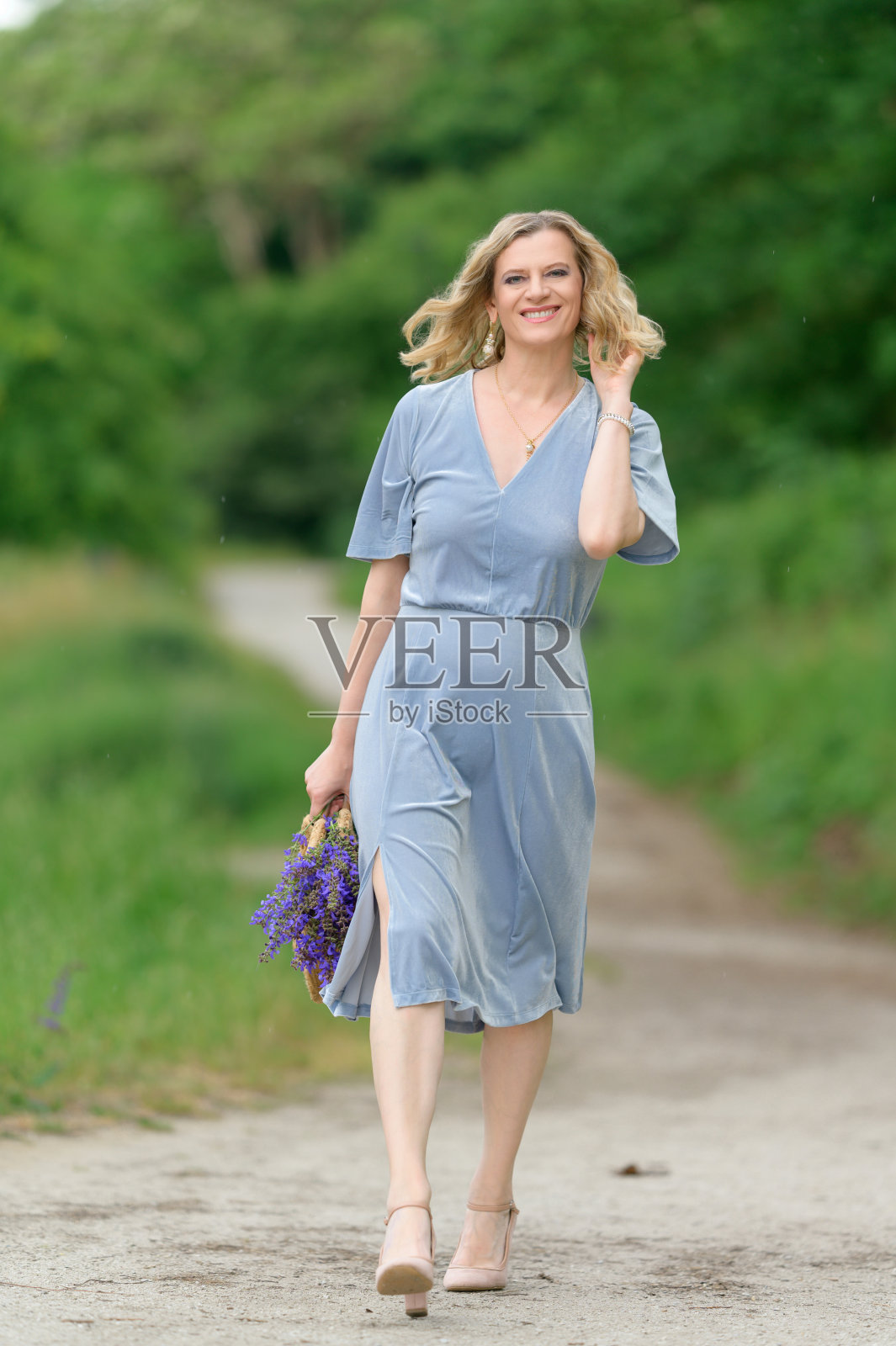 美丽的模特在春天或夏天在公园散步。蓝色的裙子,紫色的花,草编的袋子照片摄影图片