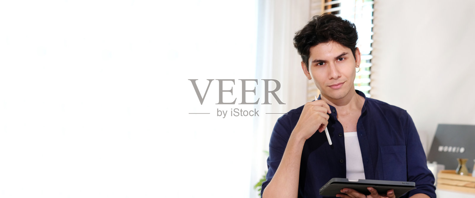 快乐的年轻商人老板的肖像拿着数码平板电脑,微笑着看着相机,而坐在家里的办公室与复制空间的背景照片摄影图片