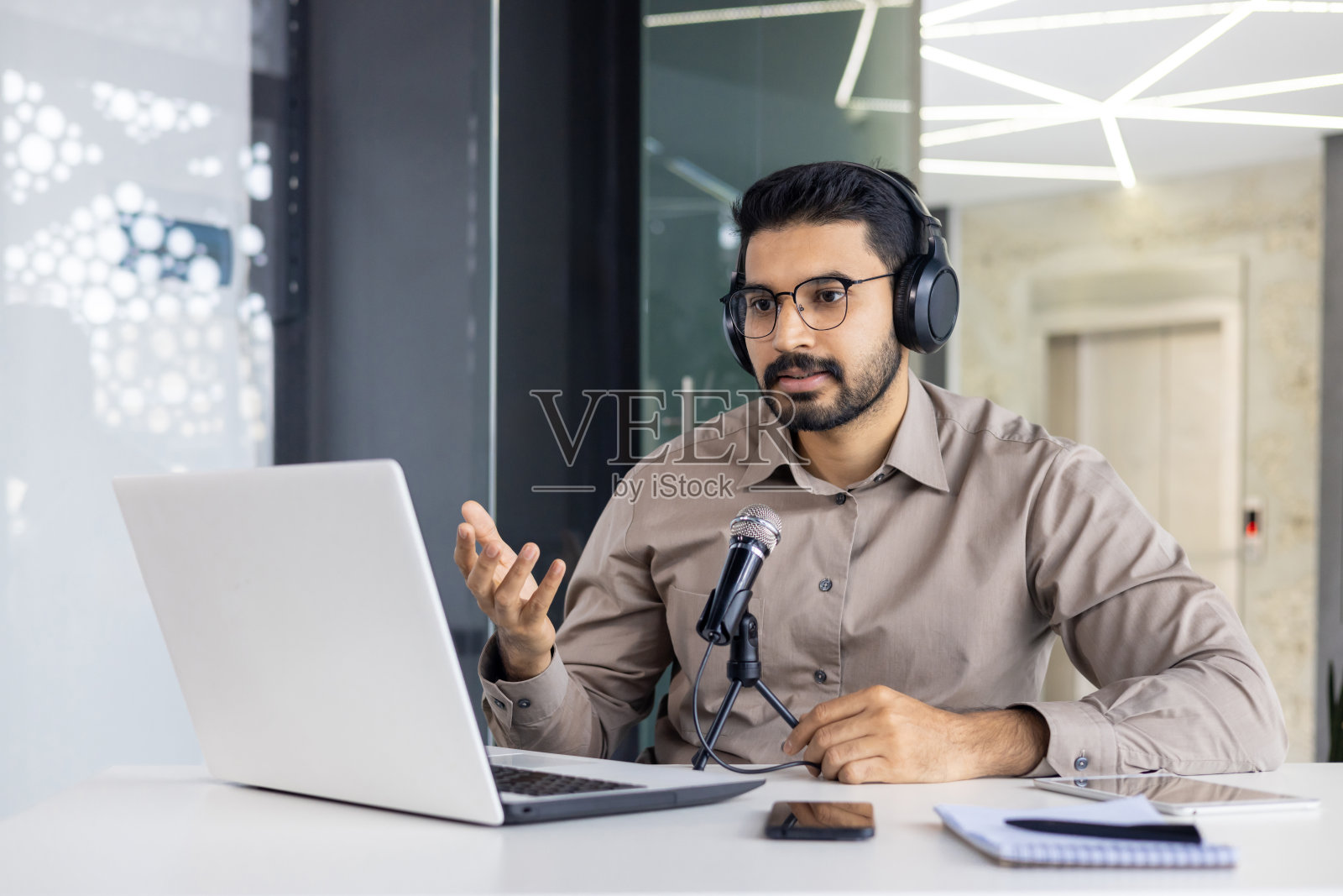 印度年轻人坐在办公室的办公桌前,戴着耳机,在笔记本电脑上对着麦克风进行视频通话,用手打手势照片摄影图片