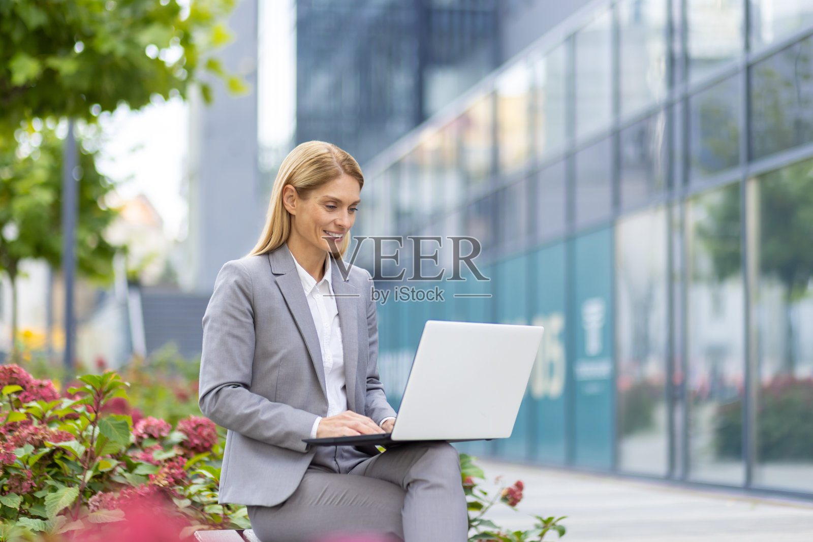 一位身穿灰色西装的年轻女子坐在办公大楼附近的长凳上,膝上放着一台笔记本电脑,正在键盘上打字照片摄影图片