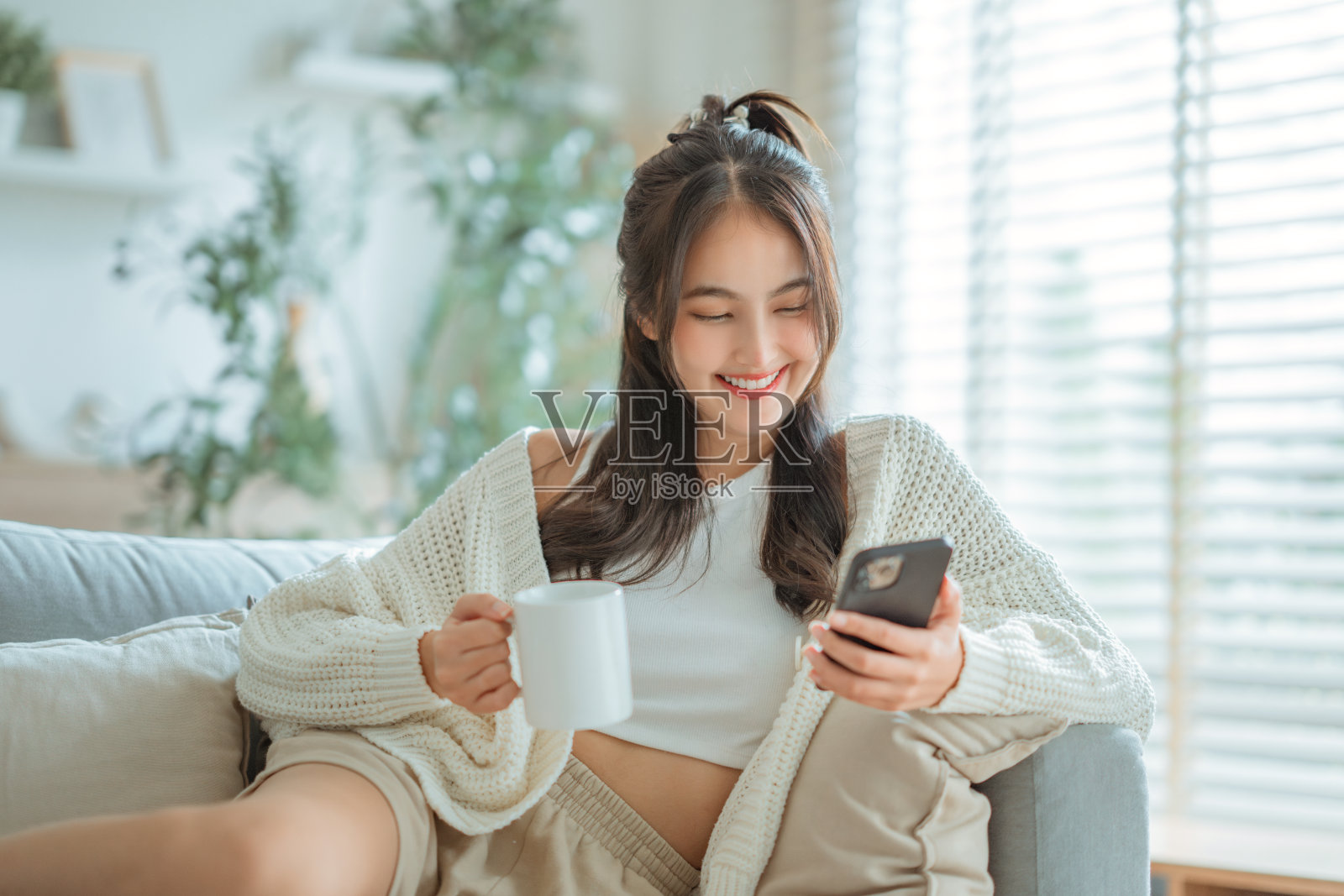 快乐的年轻亚洲女性坐在家里的沙发上,用手机上网,端着一杯咖啡或茶。照片摄影图片