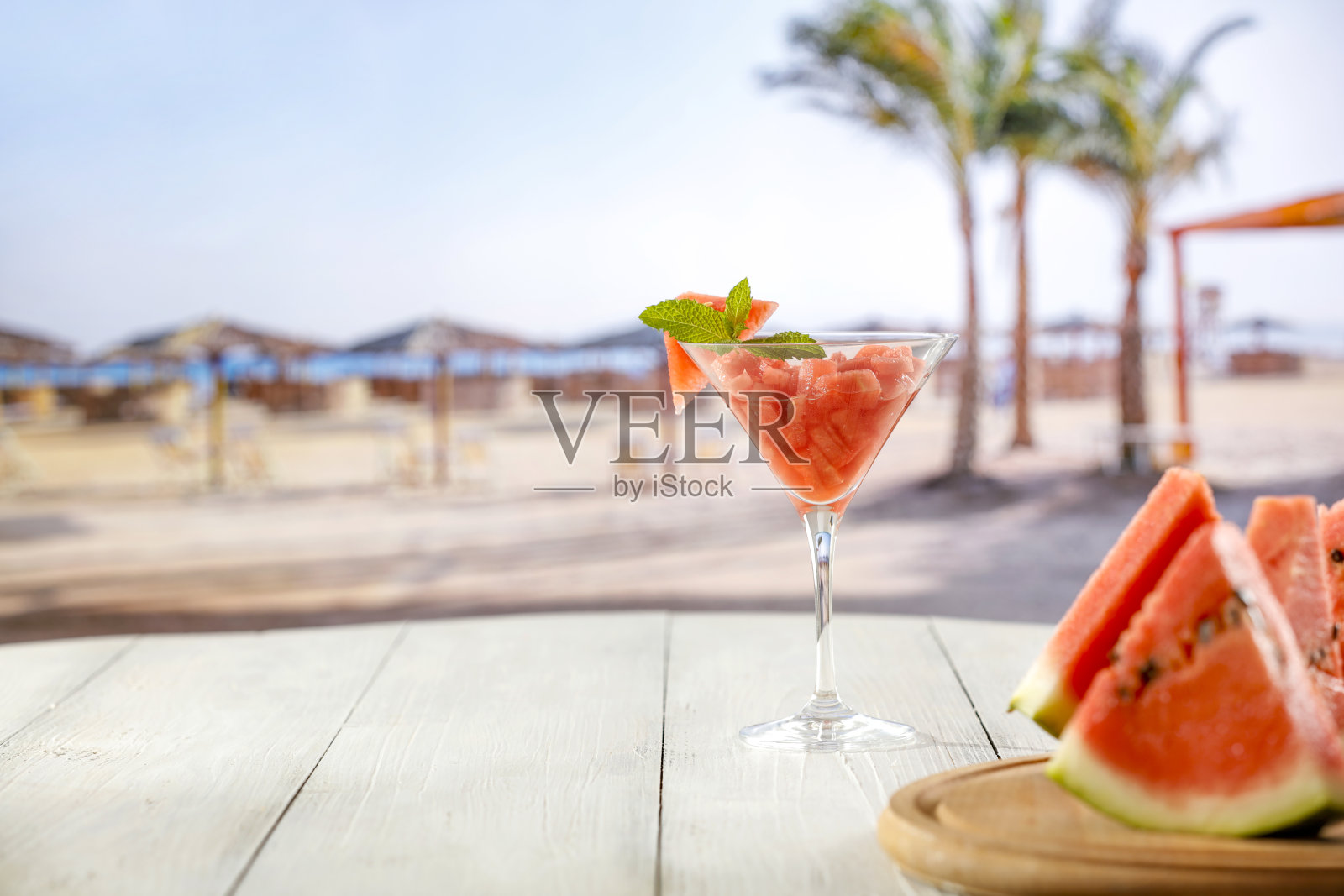 西瓜片和鸡尾酒杯在木板桌面和模糊的夏季海滩背景。照片摄影图片