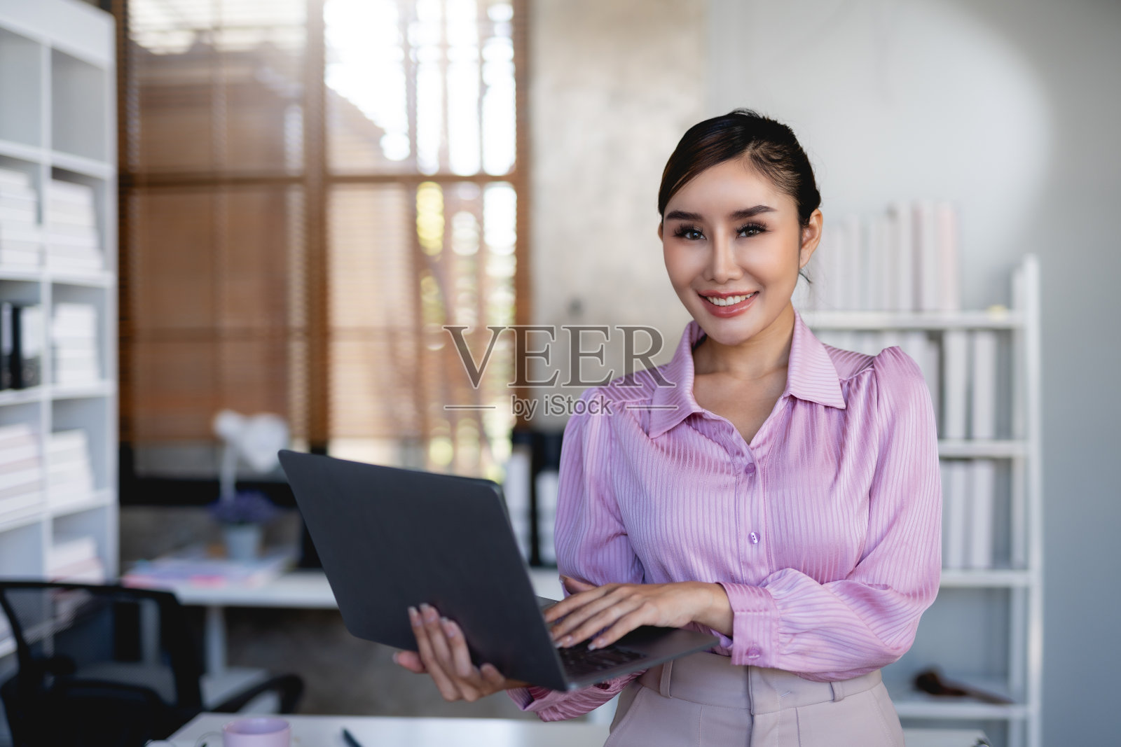 迷人微笑的亚洲女商人站在办公室拿着笔记本电脑工作。照片摄影图片