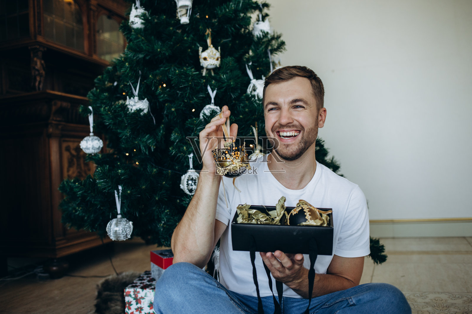 幸福英俊的年轻人在圣诞树旁拿着绿丝带的纸板箱,看着镜头。照片摄影图片
