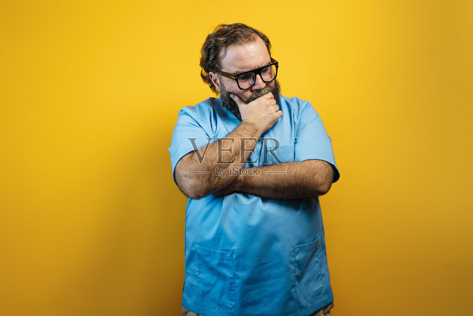 一个戴着眼镜、留着胡子的男人穿着一件蓝色衬衫,低头沉思着。他站在一个黄色的背景前。照片摄影图片