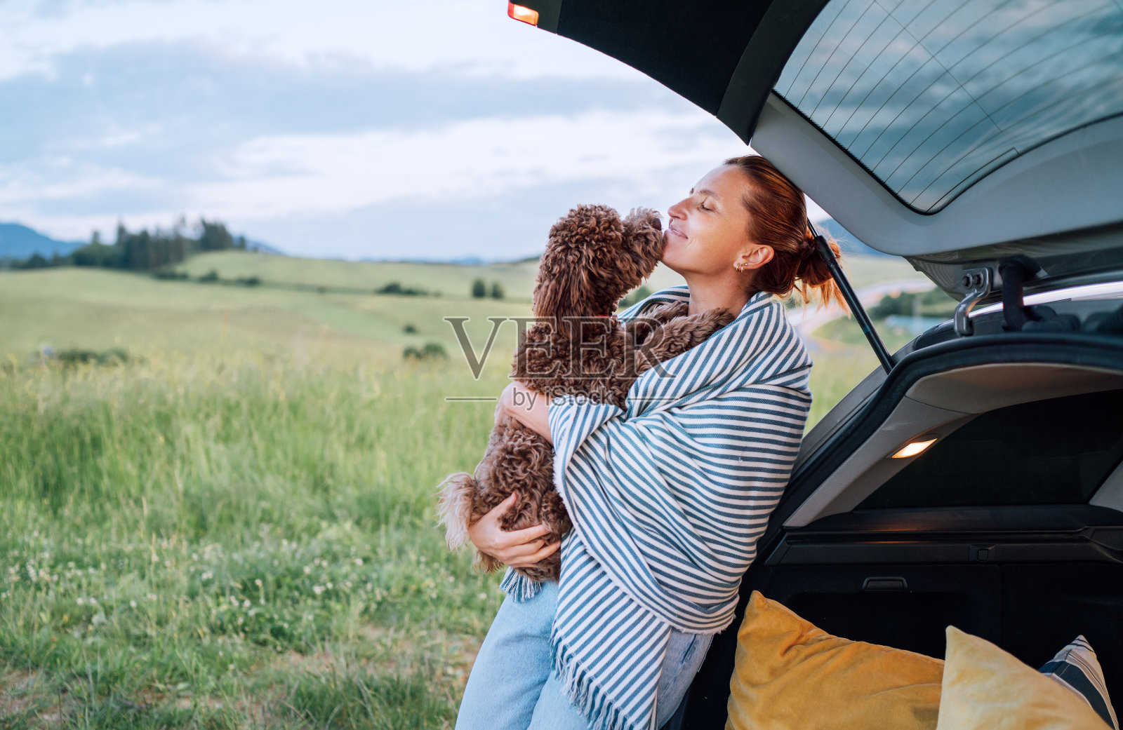 在风景如画的风景中,一个女人抱着她棕色的马提波狗,旁边是一个打开的汽车行李箱,包裹在条纹毯子里。这一场景捕捉到了宁静乡村郊游中温暖深情的瞬间照片摄影图片