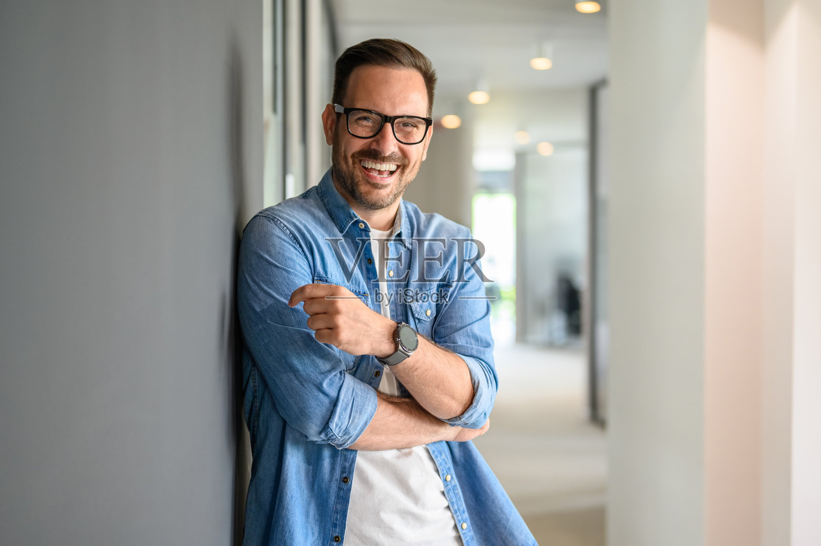 在现代办公室里,戴着眼镜笑着靠在墙上的积极的男性商业领袖的肖像照片摄影图片