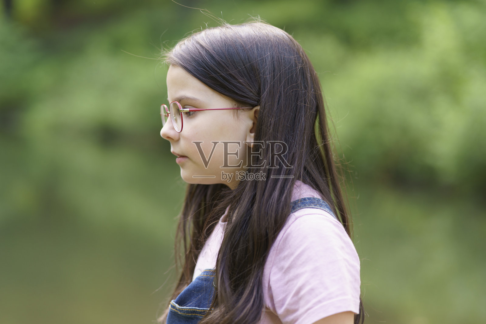 一个戴眼镜的可爱少女站在池塘边。穿着牛仔工装裤的孩子。快乐童年概念照片摄影图片