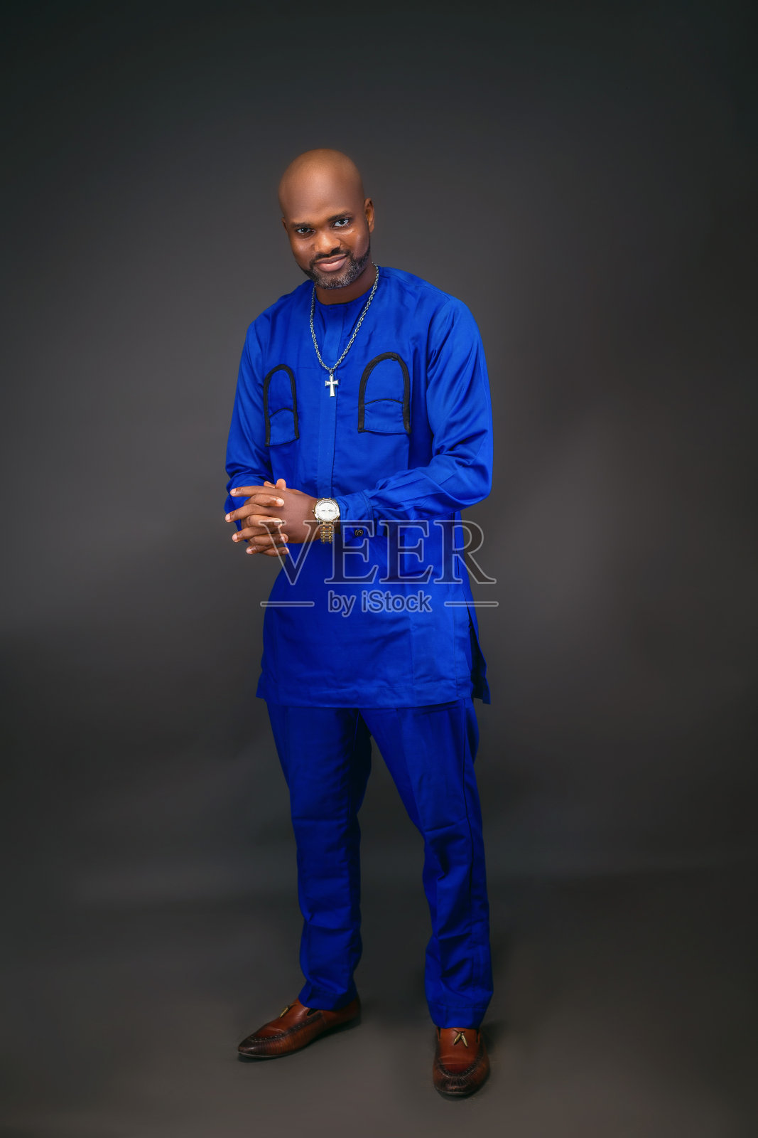 一名非洲男子身穿蓝色土著服装,戴着银链和手表,双手交叉放在棕色鞋子上,背景为浅色深色,全图照片摄影图片