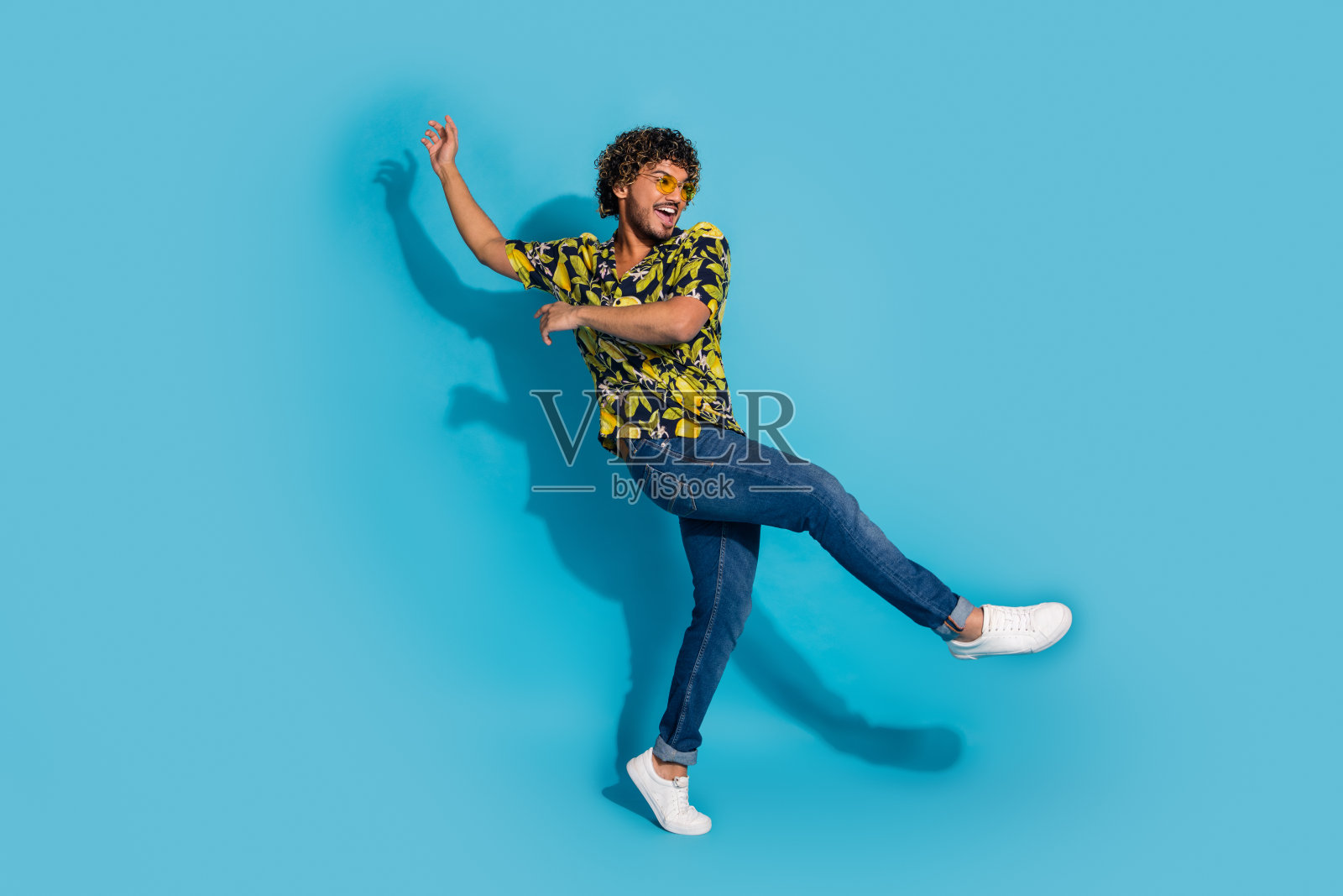疯狂的大喜过望的家伙穿着印花衬衫牛仔裤,踮着脚尖跳舞,举起手孤立在蓝色的背景照片摄影图片