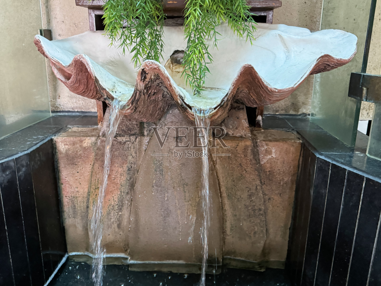 半扇贝壳设计的特写图像,室内喷泉水景,水从青铜碗倾泻到人造方形池塘,玻璃槽内填充白色鹅卵石和人造植物,重点放在前景照片摄影图片