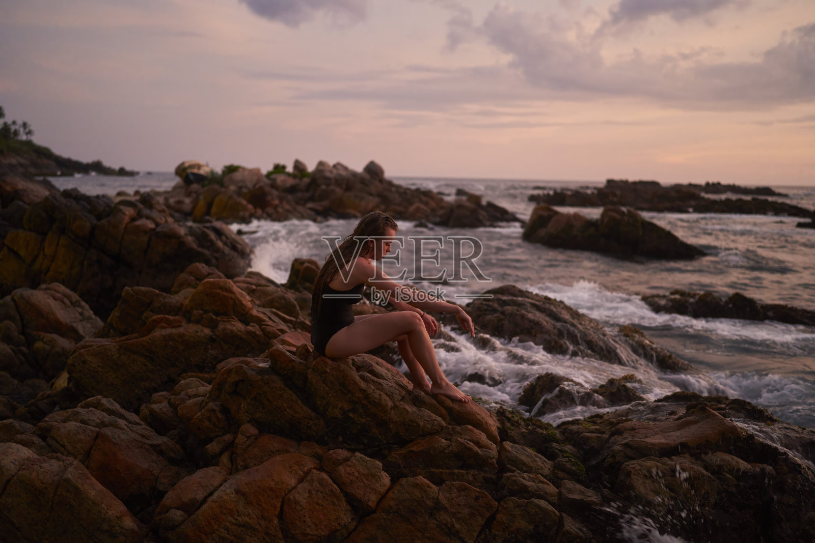 黄昏时分,女人在海边的岩石上放松,双腿浸在飞溅的海浪中,宁静的夏夜在海边。独自旅行者享受宁静的日落,与海洋景观和谐相处,健康休养。照片摄影图片