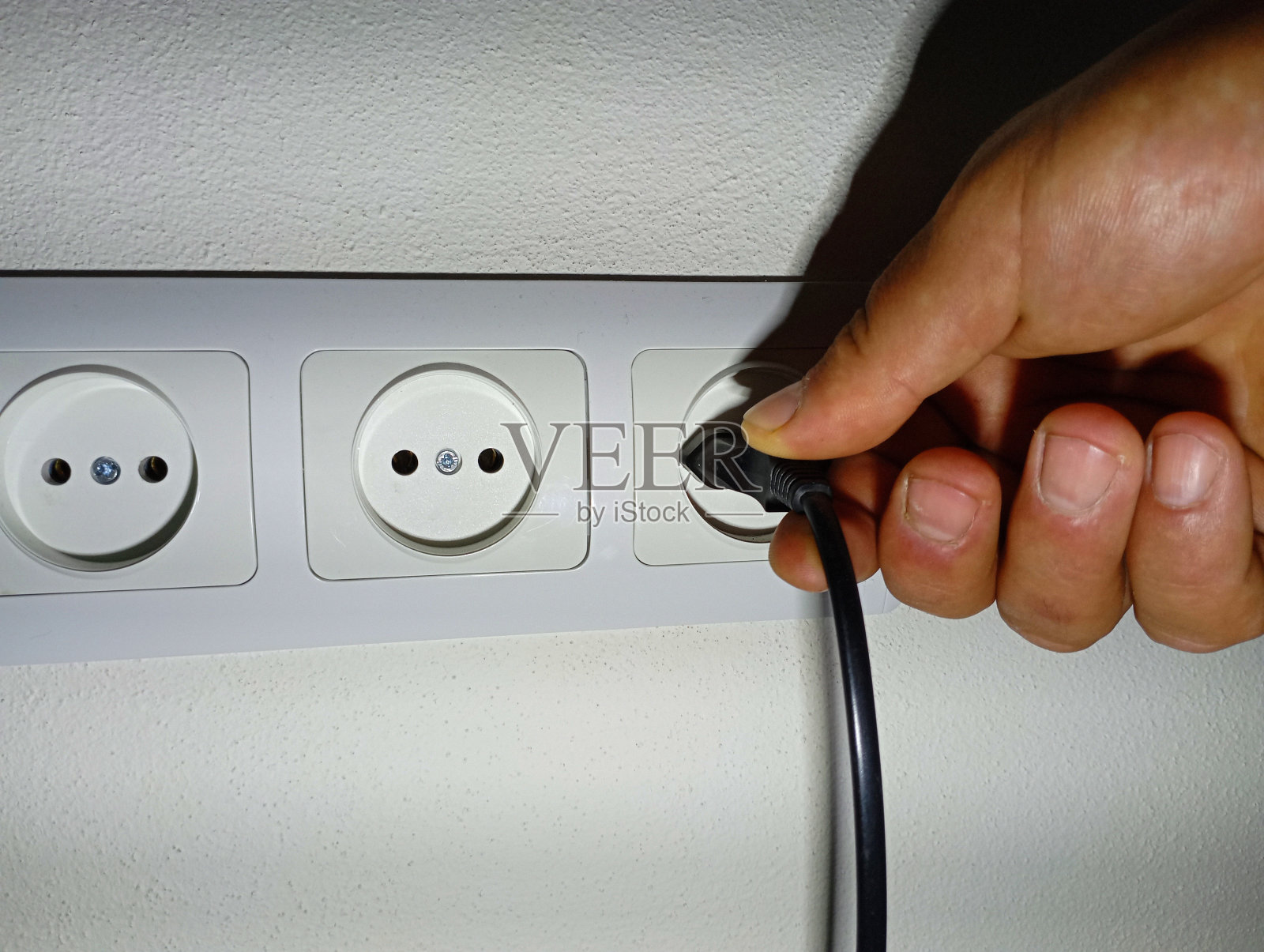 有几个插头的大电源插座,用手把设备的电源插头插进去。墙上有一个白色的电源插座。照片摄影图片