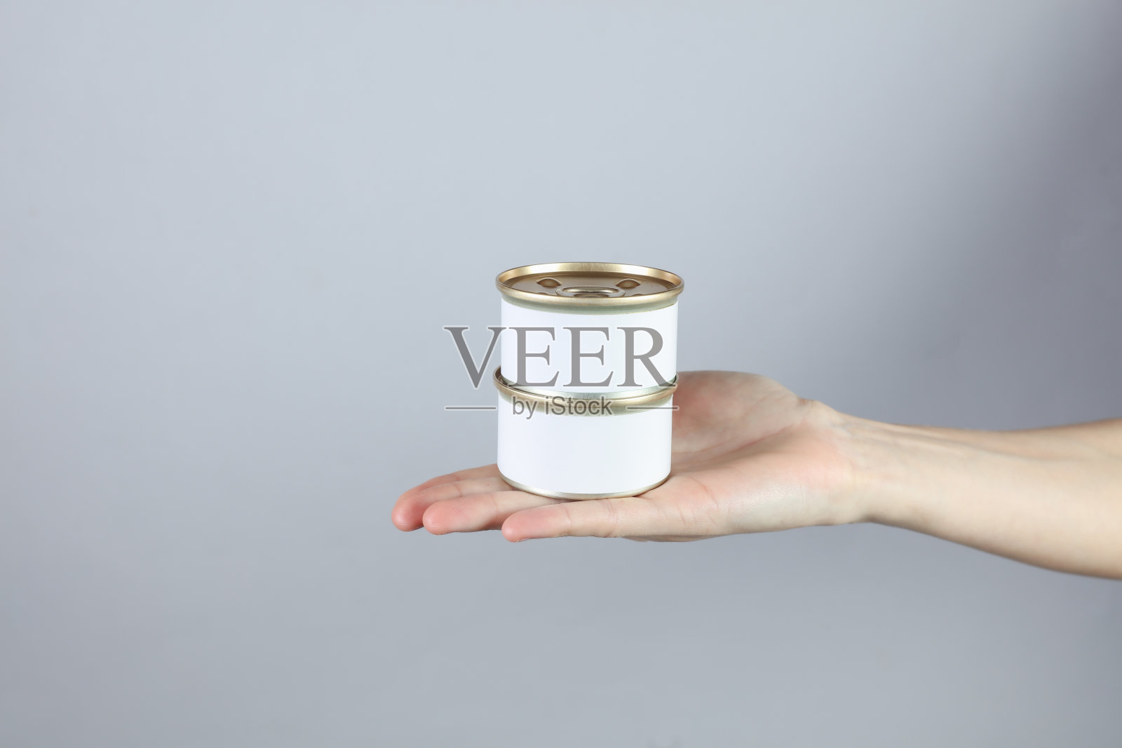 白色标签的罐头食品,一个女人的手在灰色的工作室背景上为你设计的模型照片摄影图片