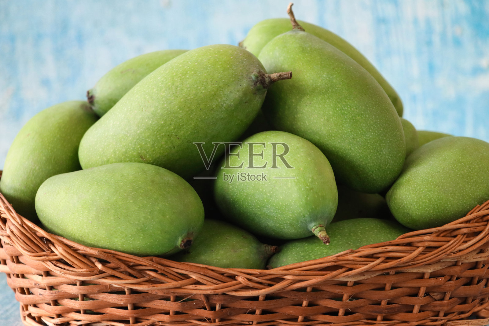 一堆完整的、生的、热带芒果水果的特写图像,绿色的皮放在柳条篮子里,蓝色和白色的背景,重点放在前景上照片摄影图片