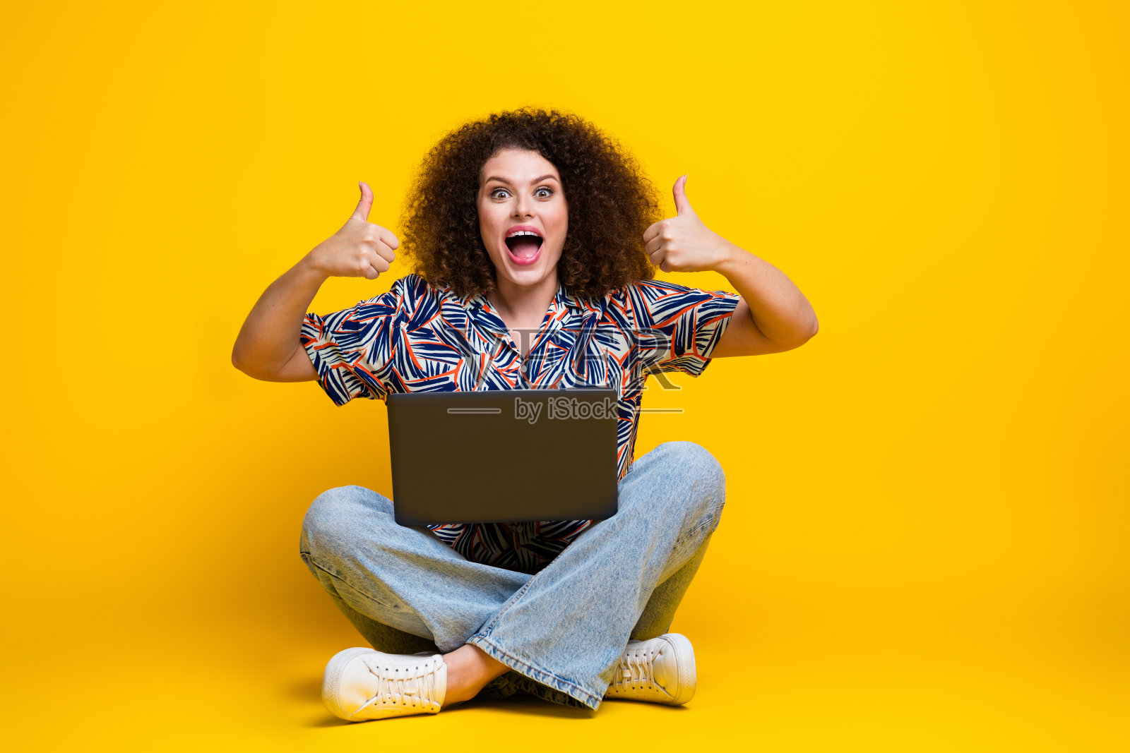 漂亮年轻女孩的全尺寸照片,坐在地板上,拿着上网本拇指向上,穿着时髦的彩色服装,孤立在黄色背景上照片摄影图片